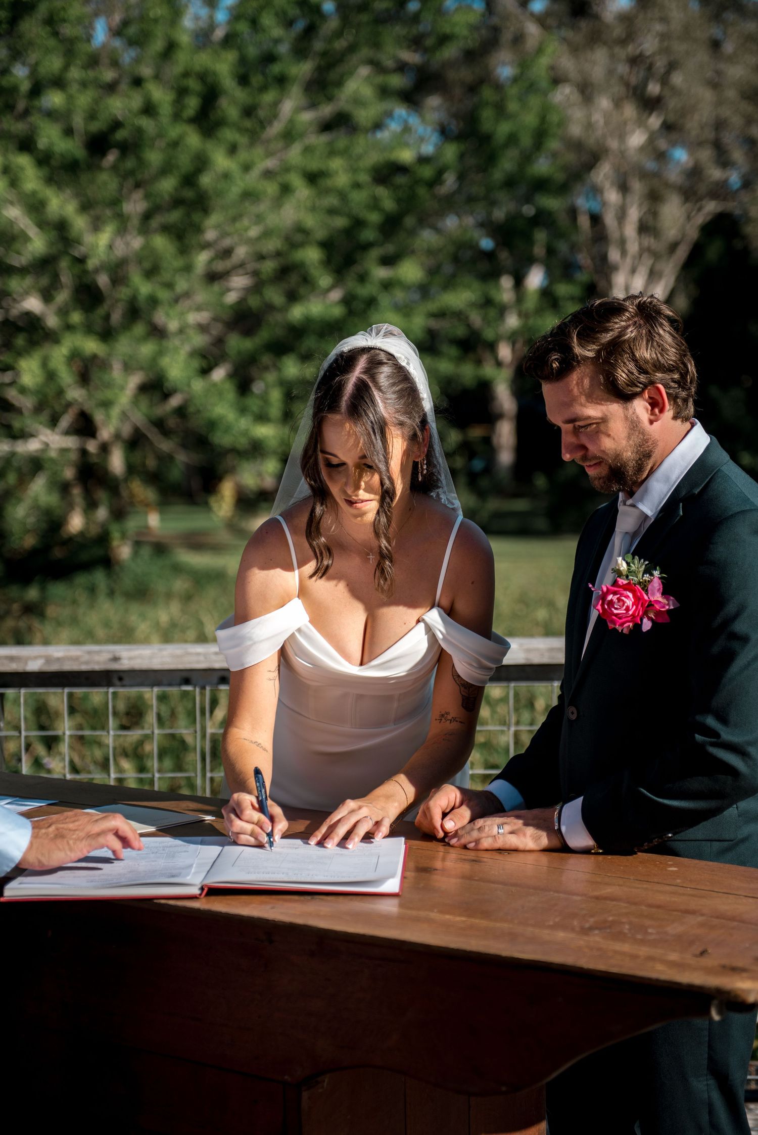 Bride signing marriage certificate with groom beside her outdoors.