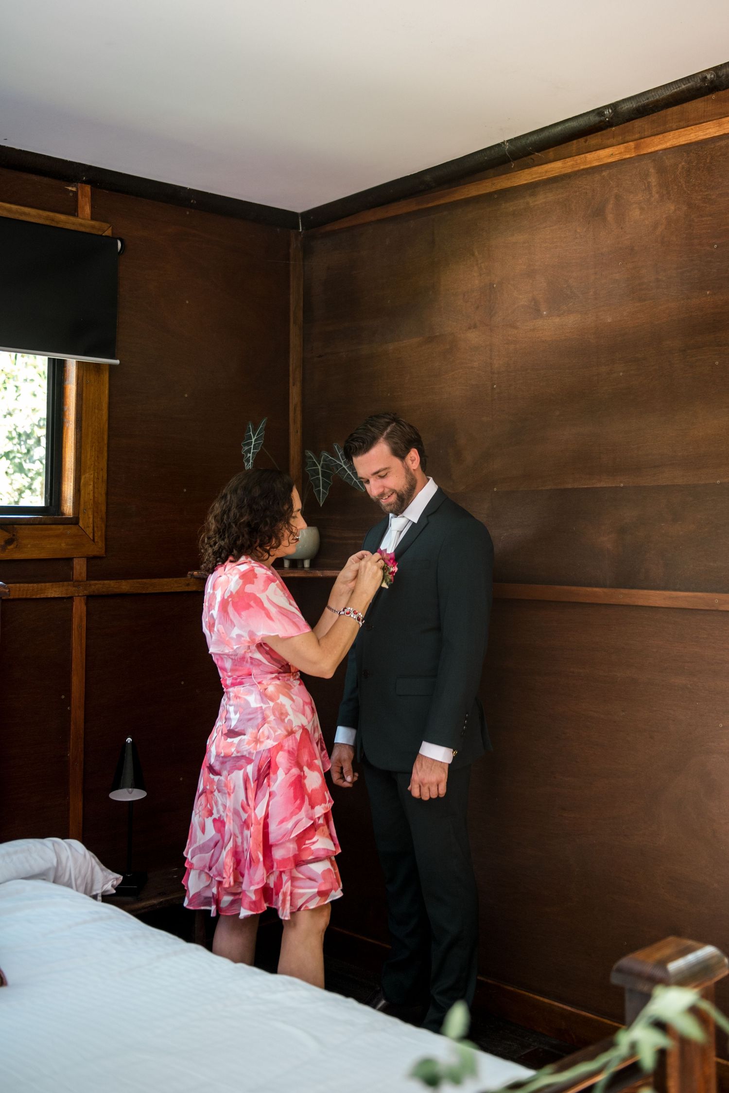 Woman in pink floral dress pinning boutonniere on groom in dark suit.