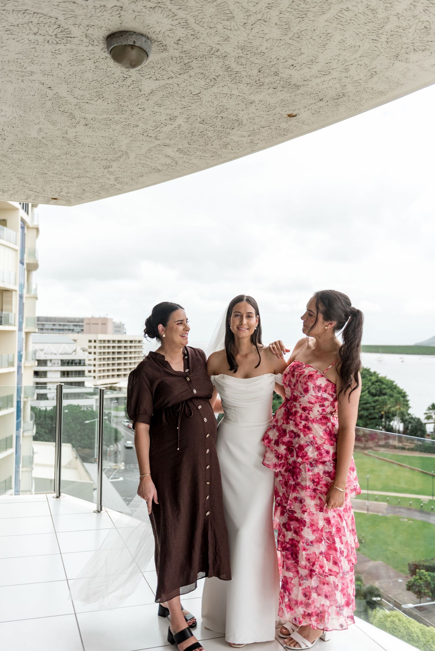 Bride in white wedding dress laughs with two guests in floral and brown dresses on balcony overlooking city.