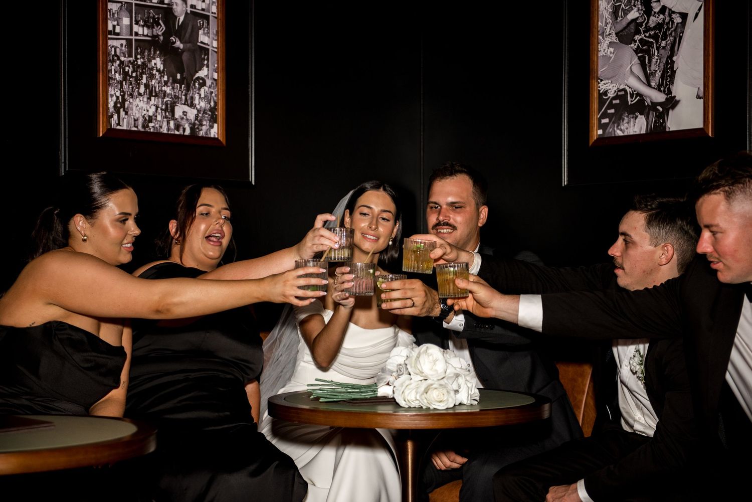 Wedding party raises glasses in a toast at a dark, intimate bar venue.