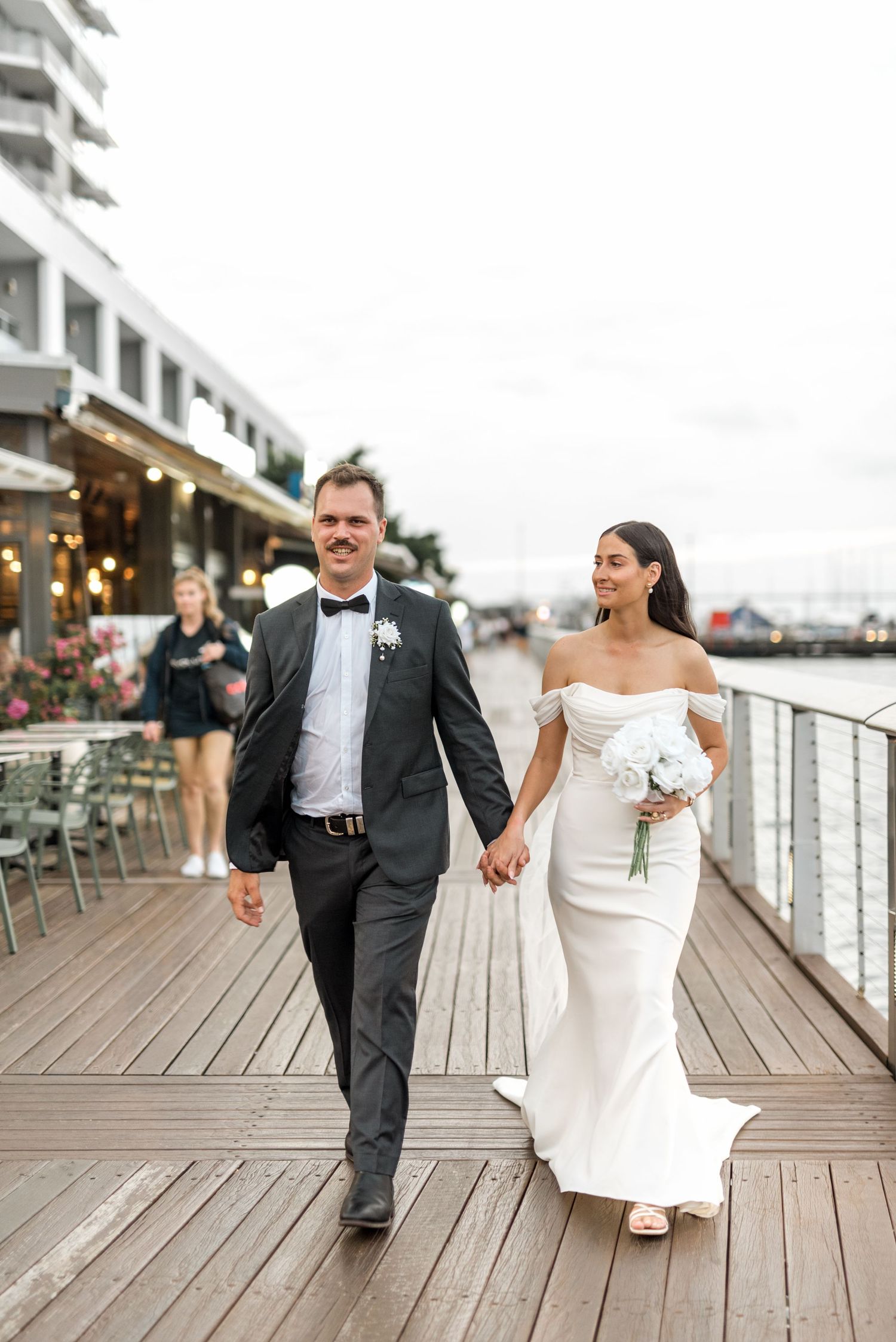 Smiling bride and groom hold hands while walking along a waterfront boardwalk after their wedding.
