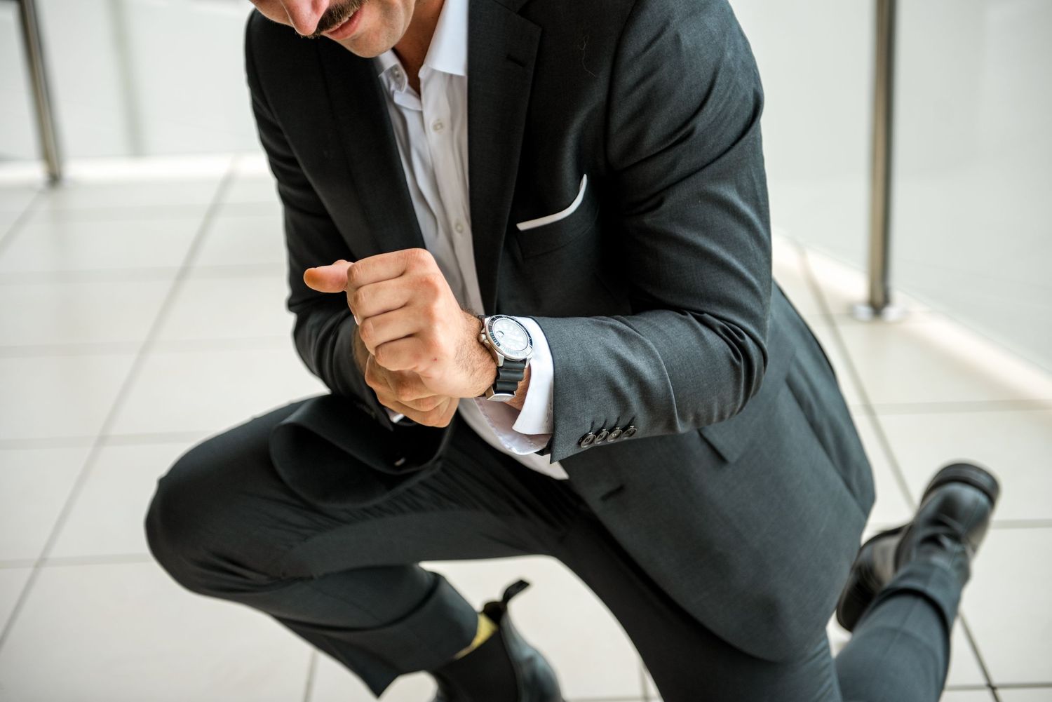 Groom in dark suit kneeling while fastening his watch cufflinks before wedding.