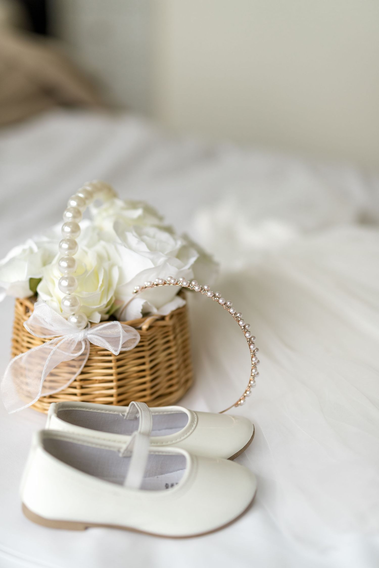 White Mary Jane flower girl shoes beside a wicker basket of white roses, pearls, and a crystal headband.