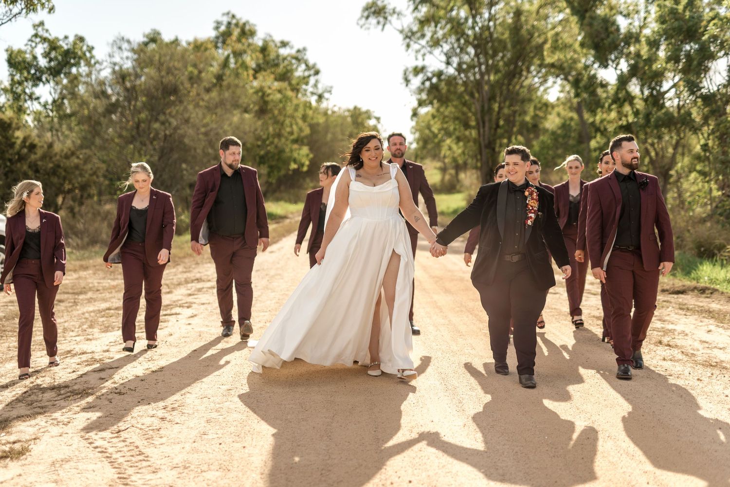 Bride and groom walk with wedding party in burgundy suits on a sandy tree-lined dirt road.