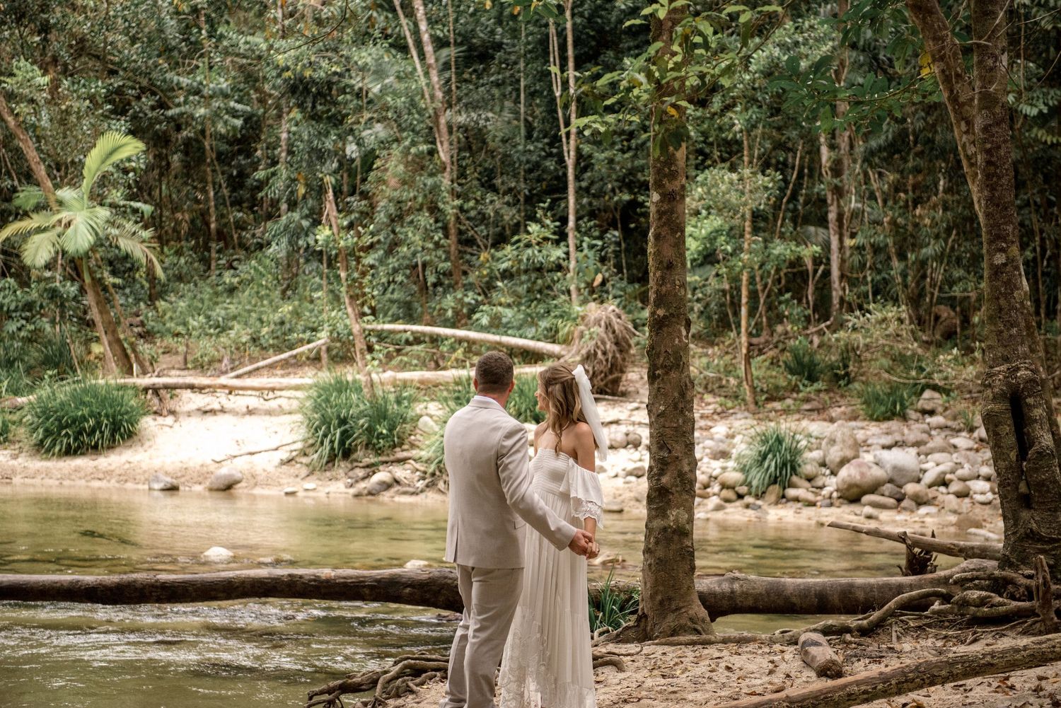 Bride and groom stand hand in hand by a serene forest creek on their wedding day.