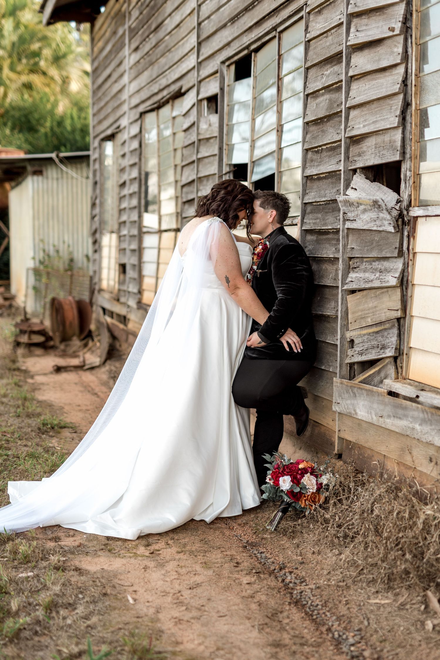 Bride and groom share a kiss against rustic wooden barn wall with bridal bouquet on ground.