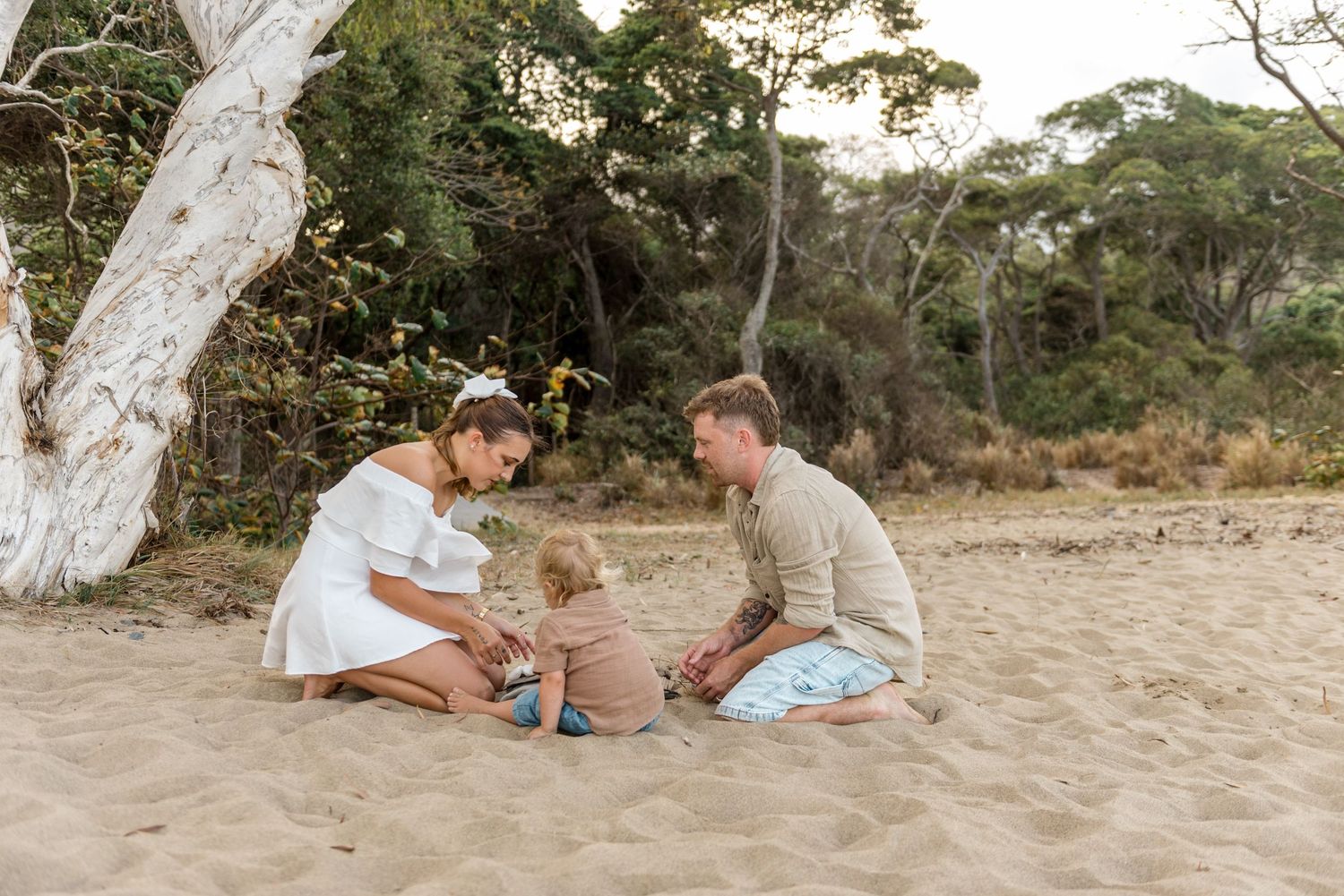 A loving family of three sits on the sand near eucalyptus trees on a peaceful beach.