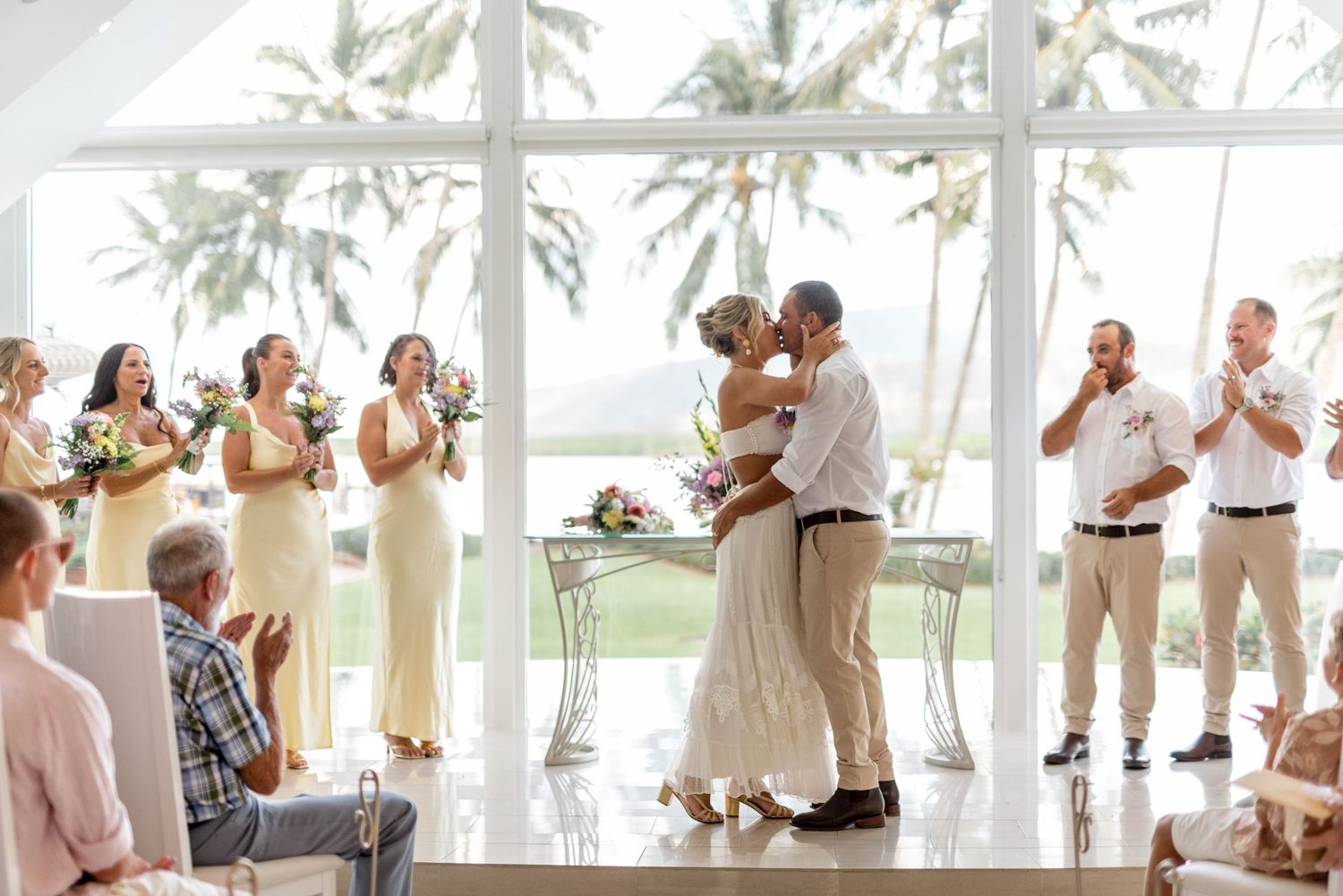 Newlyweds sharing first kiss at altar as bridal party and guests celebrate in tropical chapel.