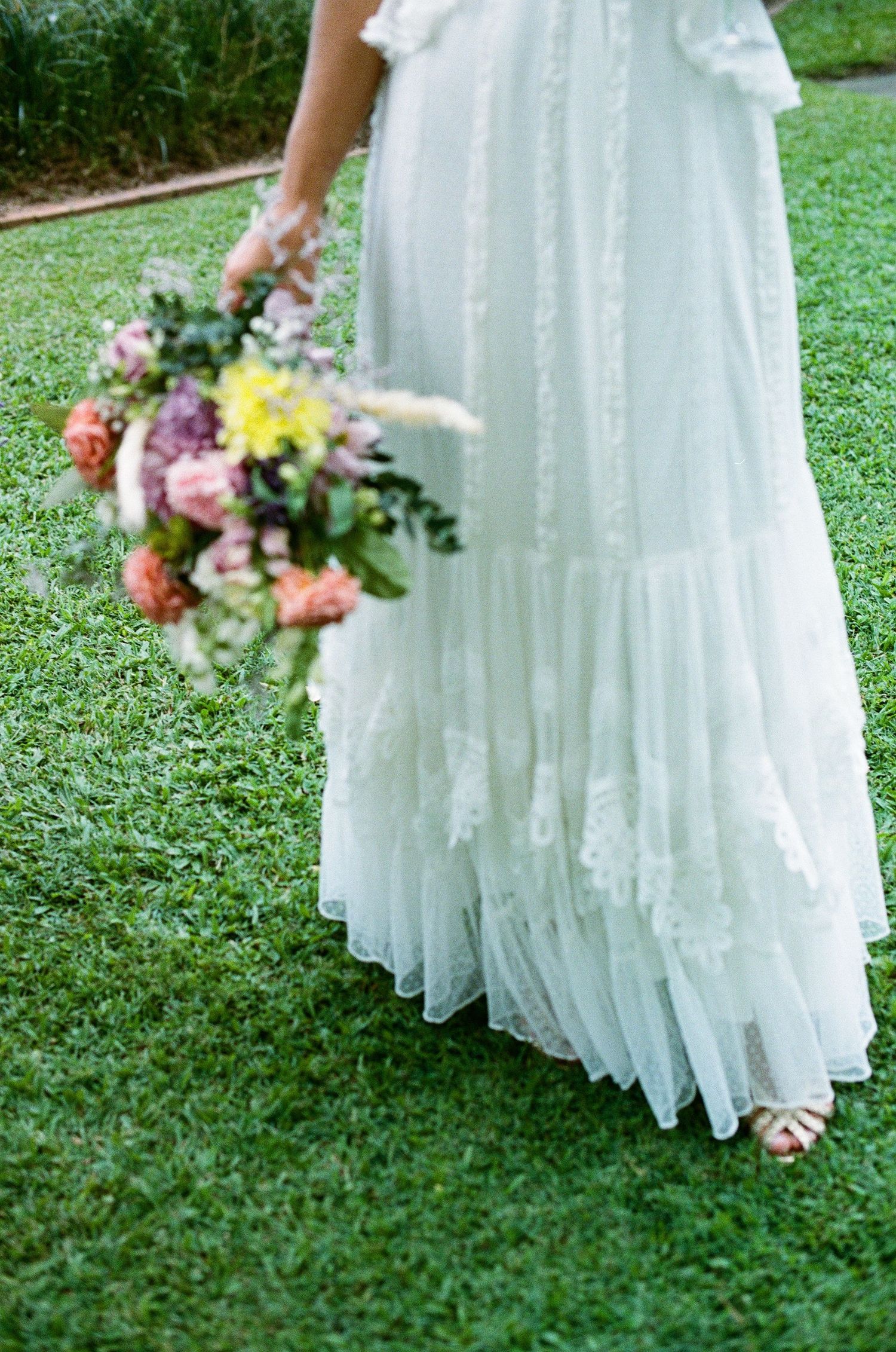 Bride in flowing white lace wedding gown holding colorful bouquet of pink, yellow, and coral flowers on green lawn.