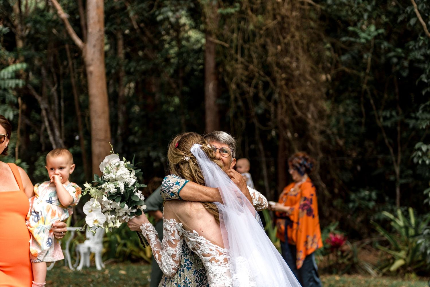 Bride hugging elderly guest while holding white bouquet after forest wedding ceremony.