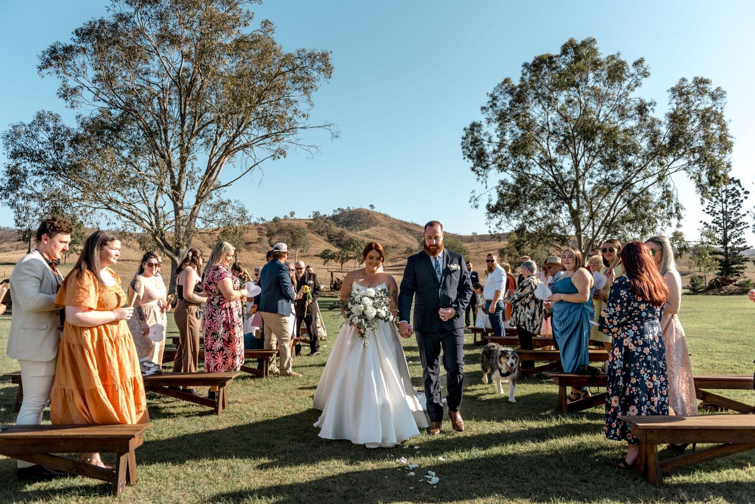 Newlyweds walk back down the aisle as guests cheer at a sunny outdoor wedding ceremony on green grass.