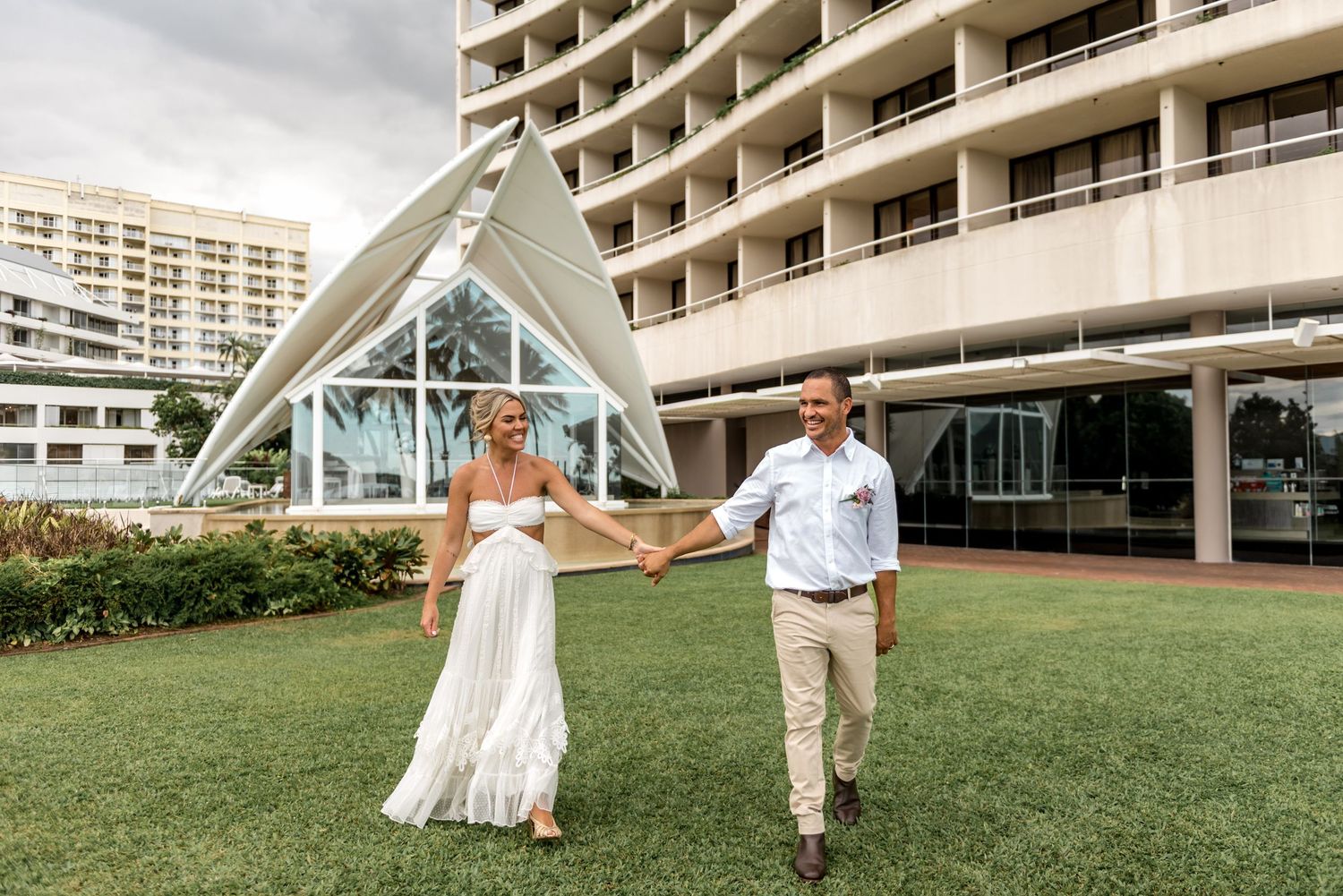 Smiling bride and groom walk hand in hand on green lawn outside modern hotel with glass chapel.