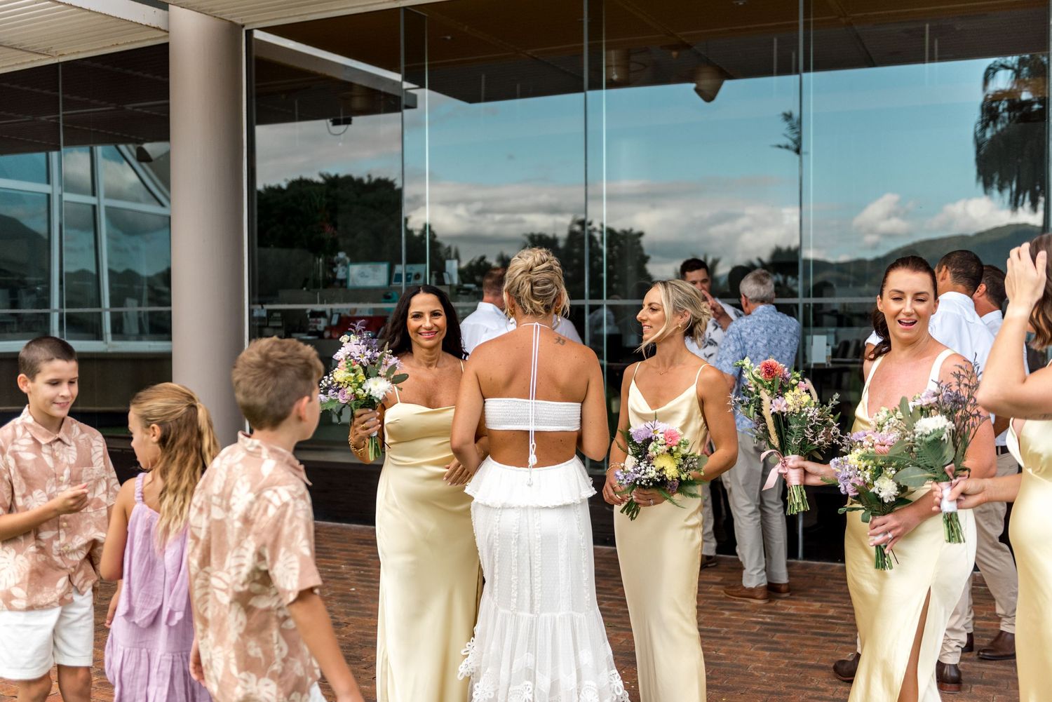 Bride and bridesmaids in yellow dresses mingle with wedding guests outside a modern glass building.