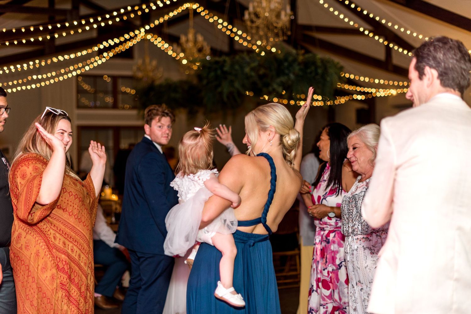Wedding guests dance together on the reception floor under warm string lights in a rustic venue.