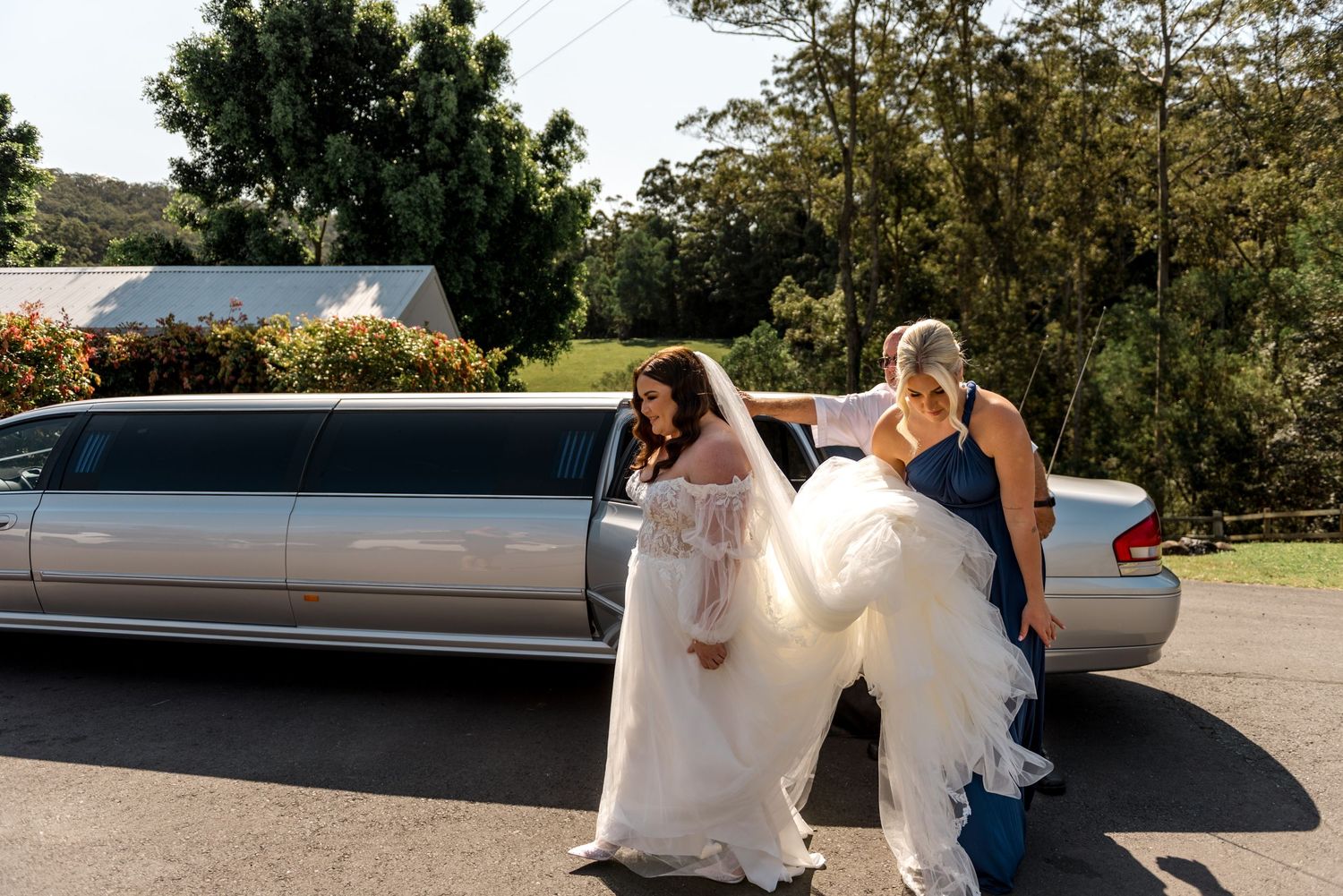Bride in a lace wedding gown being helped by bridesmaid exiting a white stretch limousine outdoors.