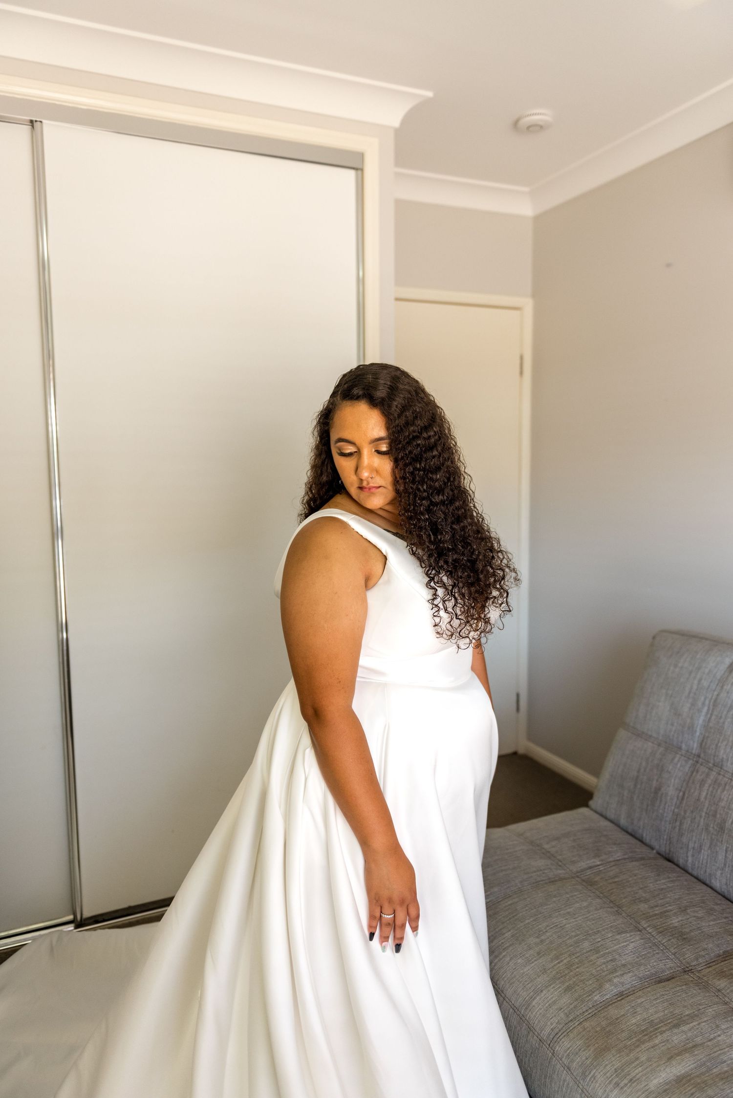 Bride in elegant white ball gown with curly hair standing in bedroom.