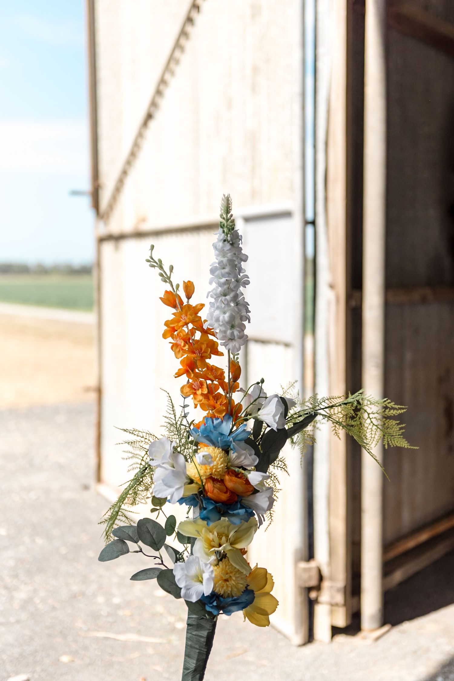 Colorful bridal bouquet with orange, white, and blue flowers outside a rustic barn.