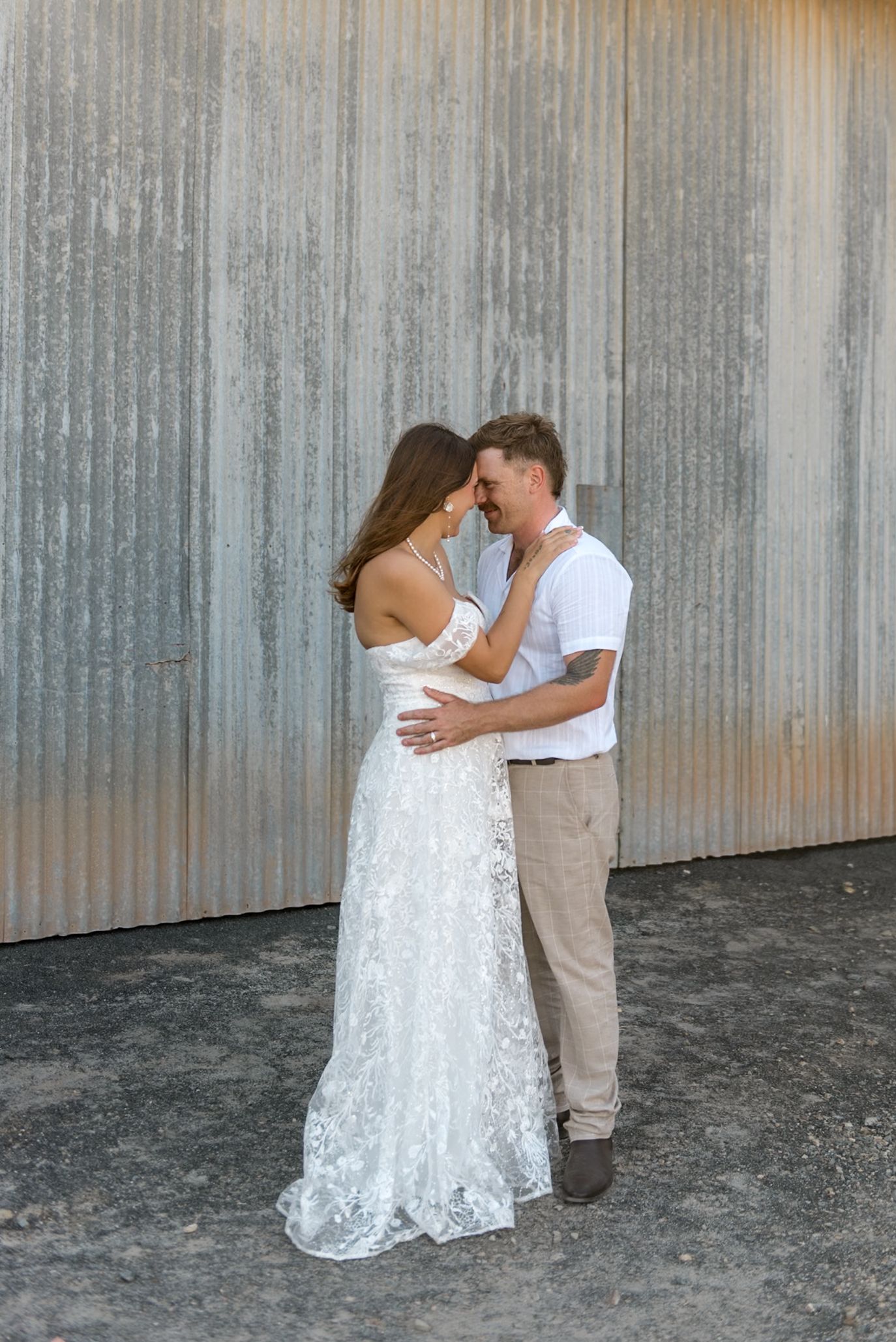 Bride in lace gown and groom in white shirt embrace tenderly in front of rustic corrugated metal wall.