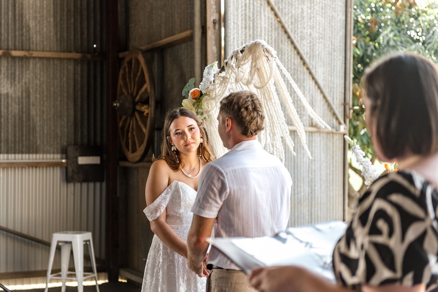 Smiling bride in white lace dress holds grooms hands during intimate barn wedding ceremony with macrame arch.