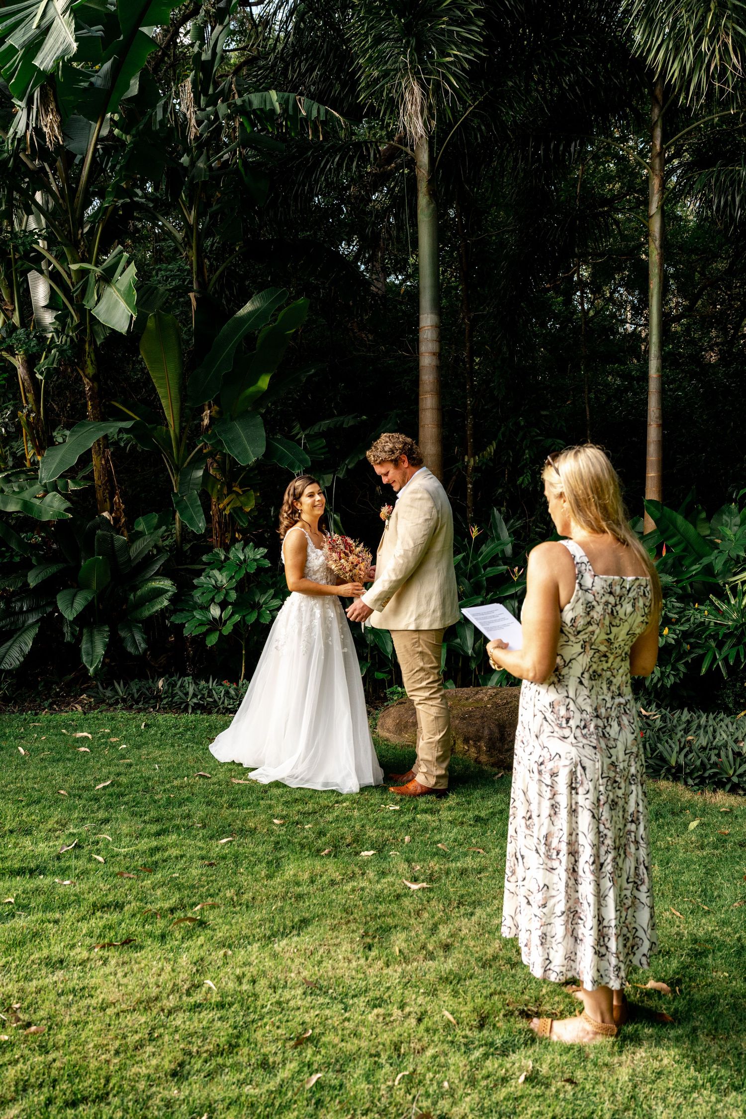 A bride and groom exchange vows outdoors in a tropical garden with a celebrant nearby.