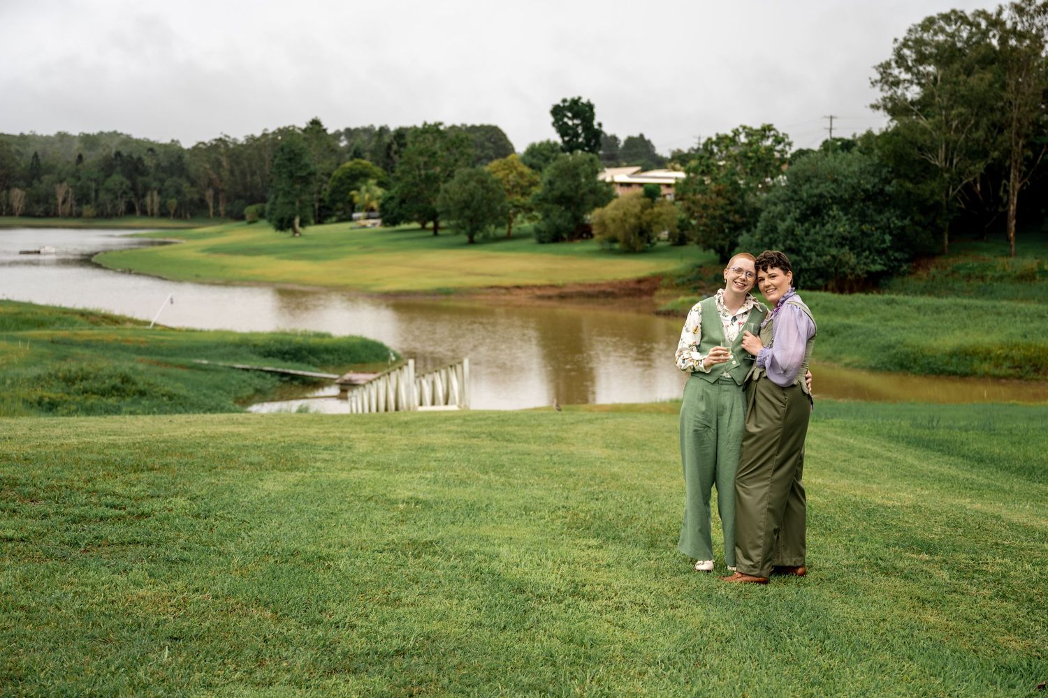 Two brides embracing on green lawn with scenic pond and trees in background.