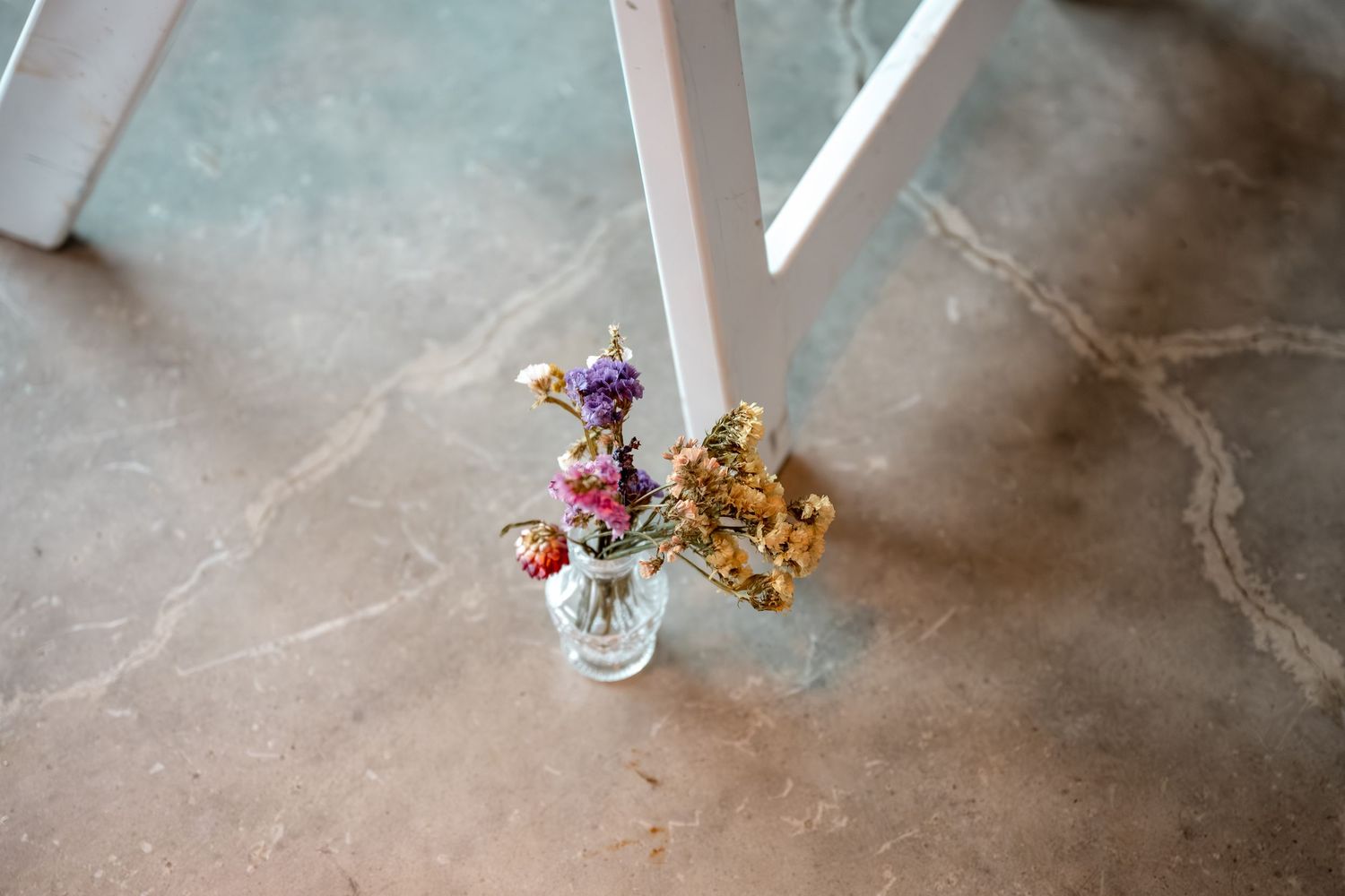 Small glass vase with colorful dried wildflowers beside a white chair leg on a concrete floor.