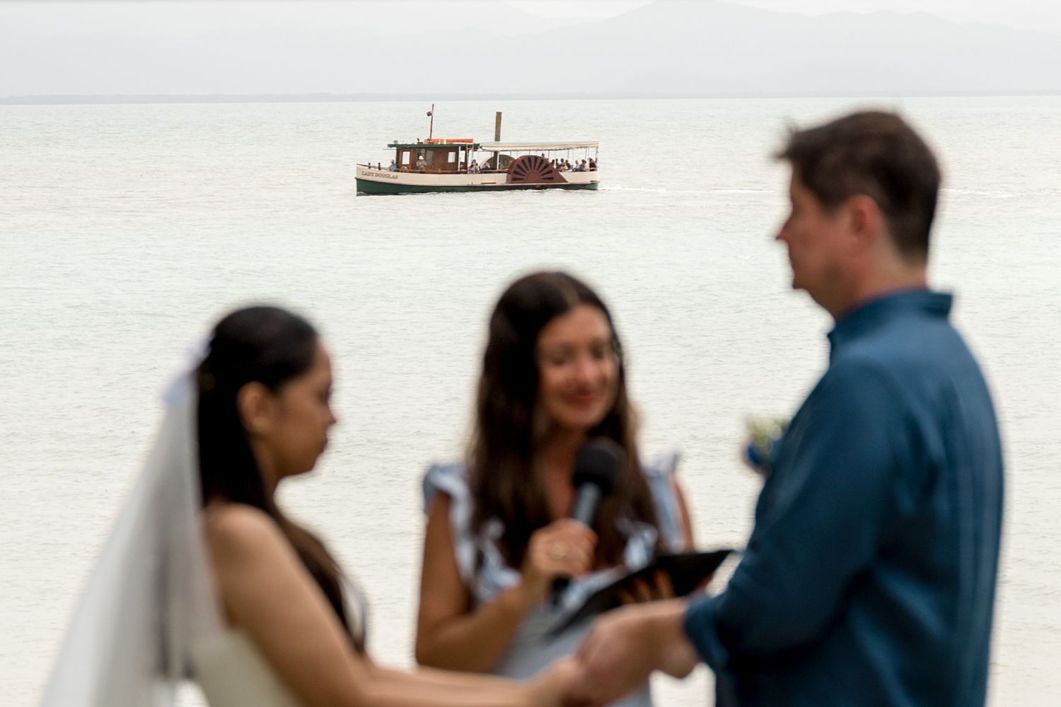 Bride and groom exchange vows with officiant as a vintage wooden boat floats on calm ocean waters behind at Little Cove, Port Douglas.