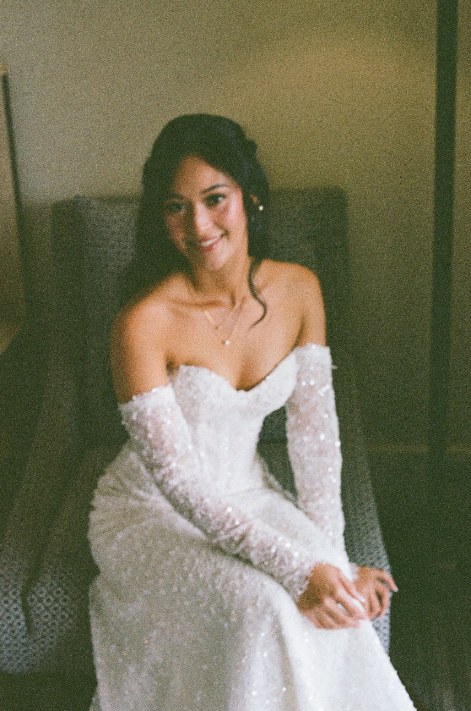 Radiant bride in beaded off-shoulder wedding gown seated in chair smiling.