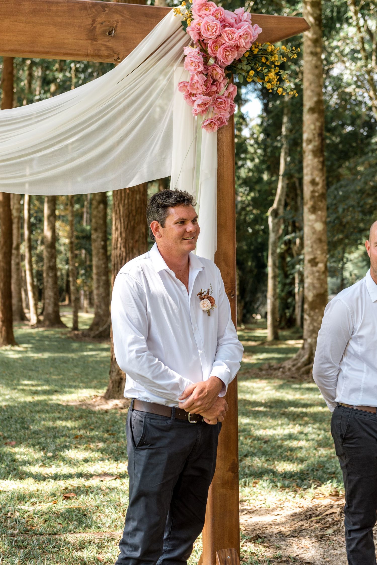 Smiling groom in white shirt stands beneath a decorated wooden arch with pink roses.