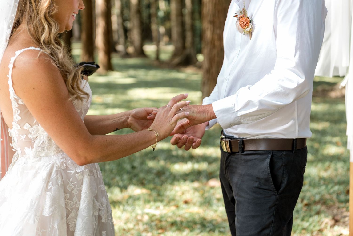 Bride places wedding ring on grooms finger during outdoor forest wedding ceremony exchange.