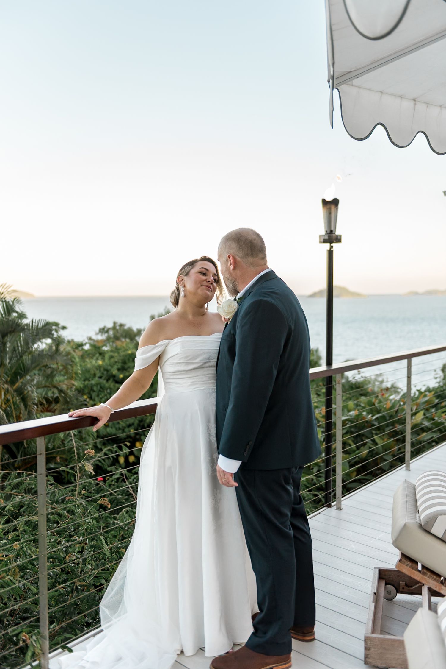 Bride in off-shoulder gown and groom share an intimate moment on a balcony overlooking tropical ocean scenery.