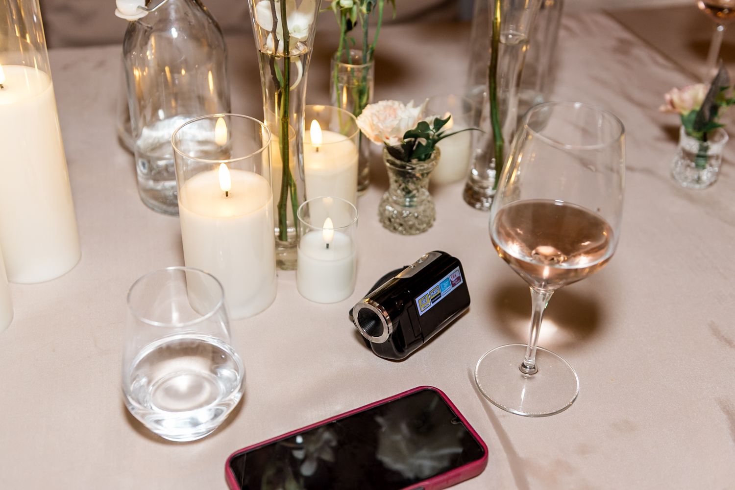 Wedding reception table with candles, wine glasses, a small video camera, and a pink phone on white tablecloth.