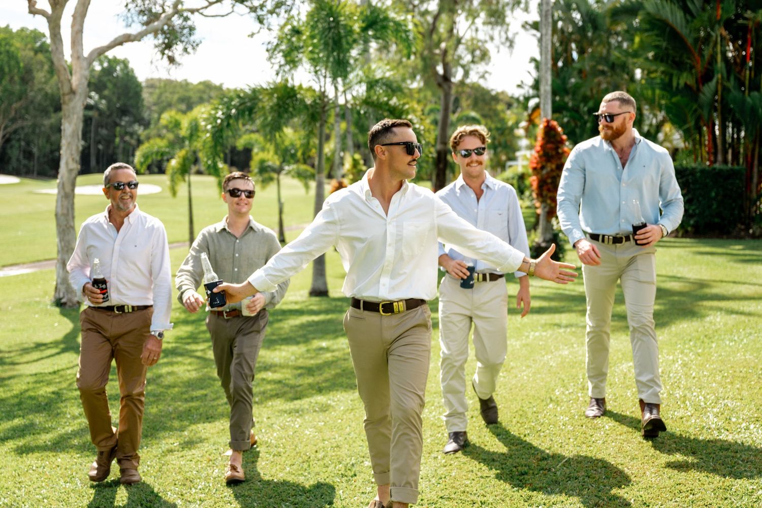 Groomsmen in white shirts and khaki pants walking outdoors with one spreading arms wide on grass.