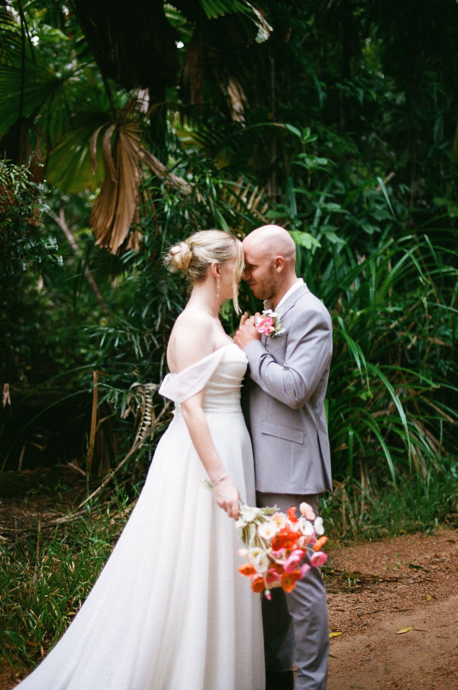 Bride and groom sharing an intimate moment surrounded by lush tropical greenery, bride holding a vibrant coral bouquet.