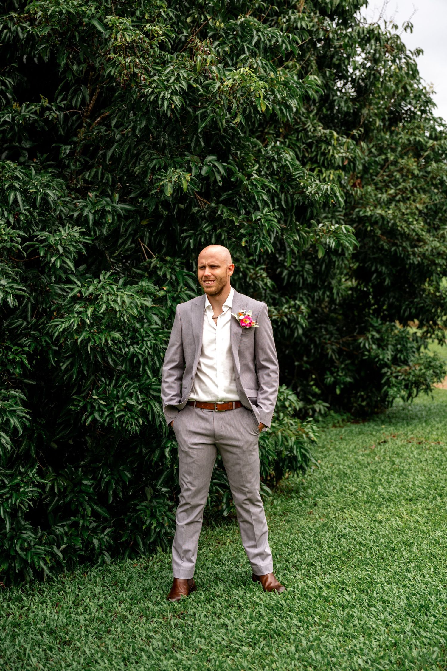 Smiling bald groom in grey suit with pink boutonniere standing on green lawn before trees.