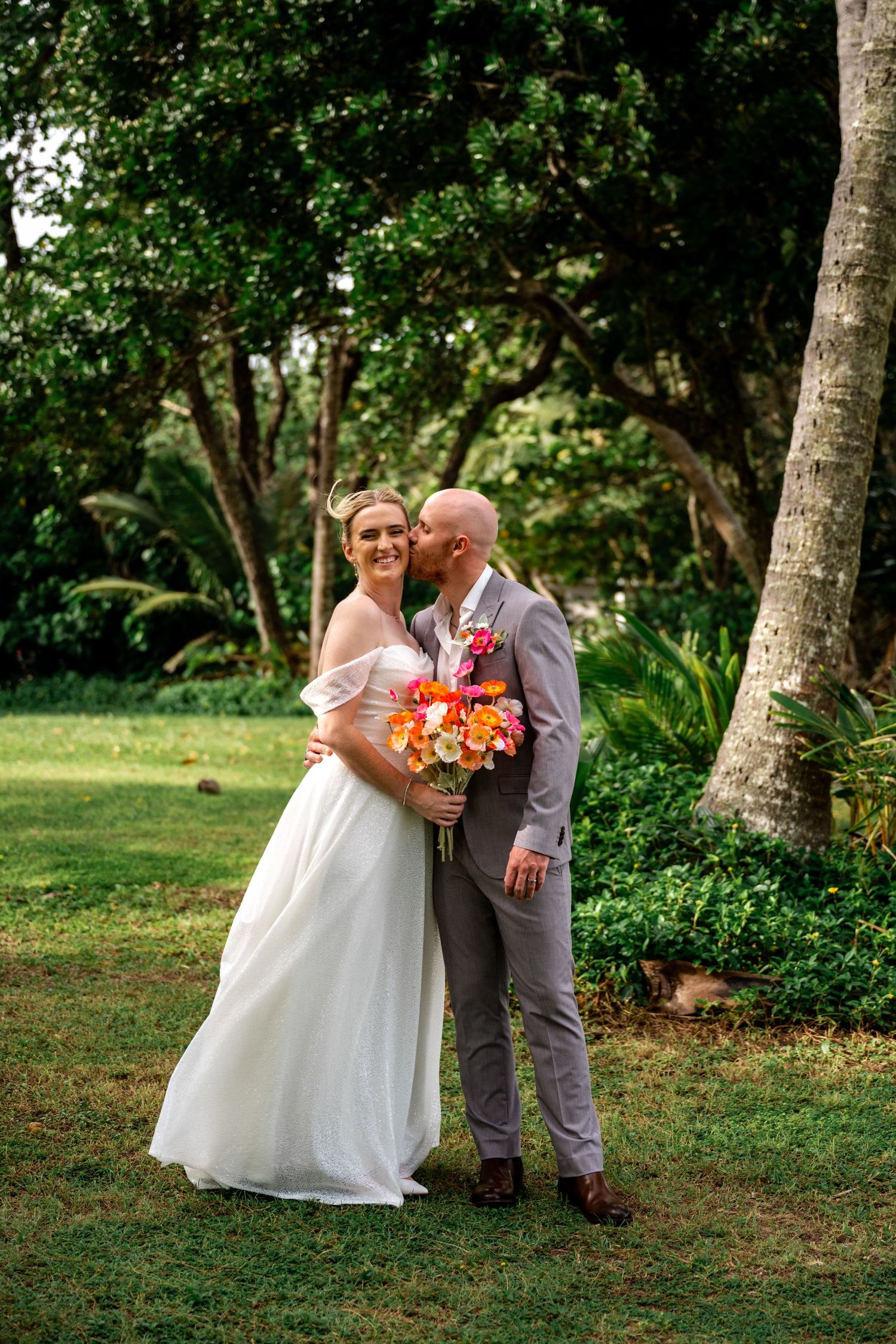 Groom in grey suit kissing bride on cheek while she smiles holding bright tropical bouquet in garden setting.