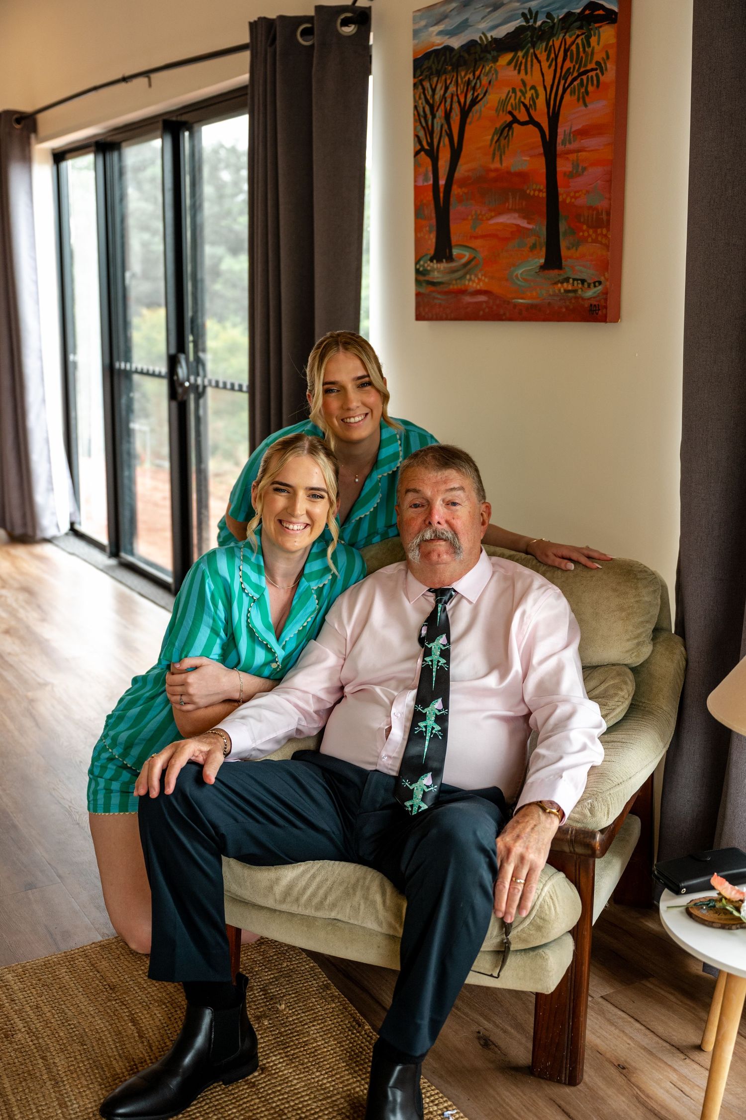 Bride in teal robe with older man and bridesmaid posing together in living room before wedding.