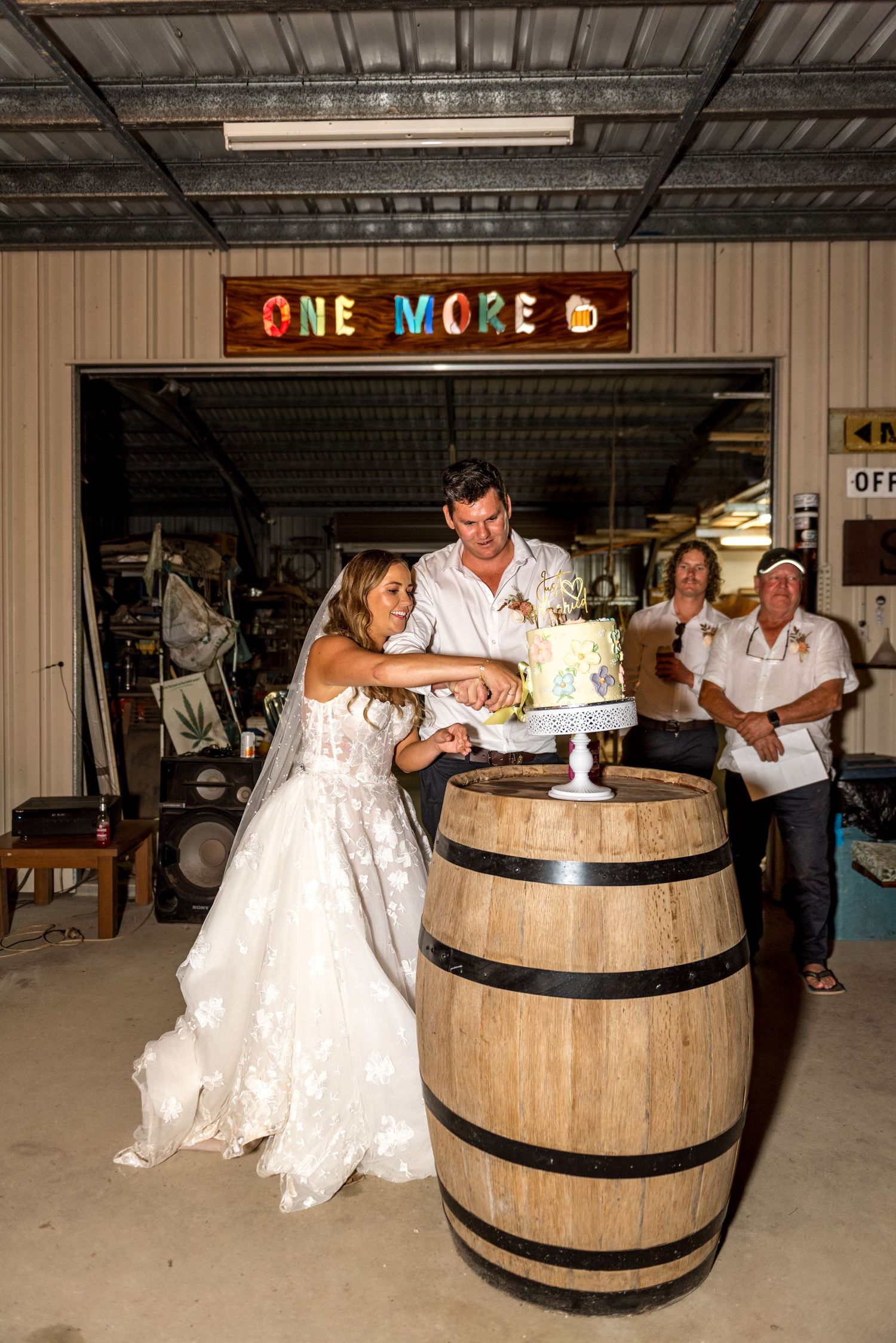 Bride and groom cutting wedding cake on rustic wooden barrel inside shed with One More sign and guests watching.