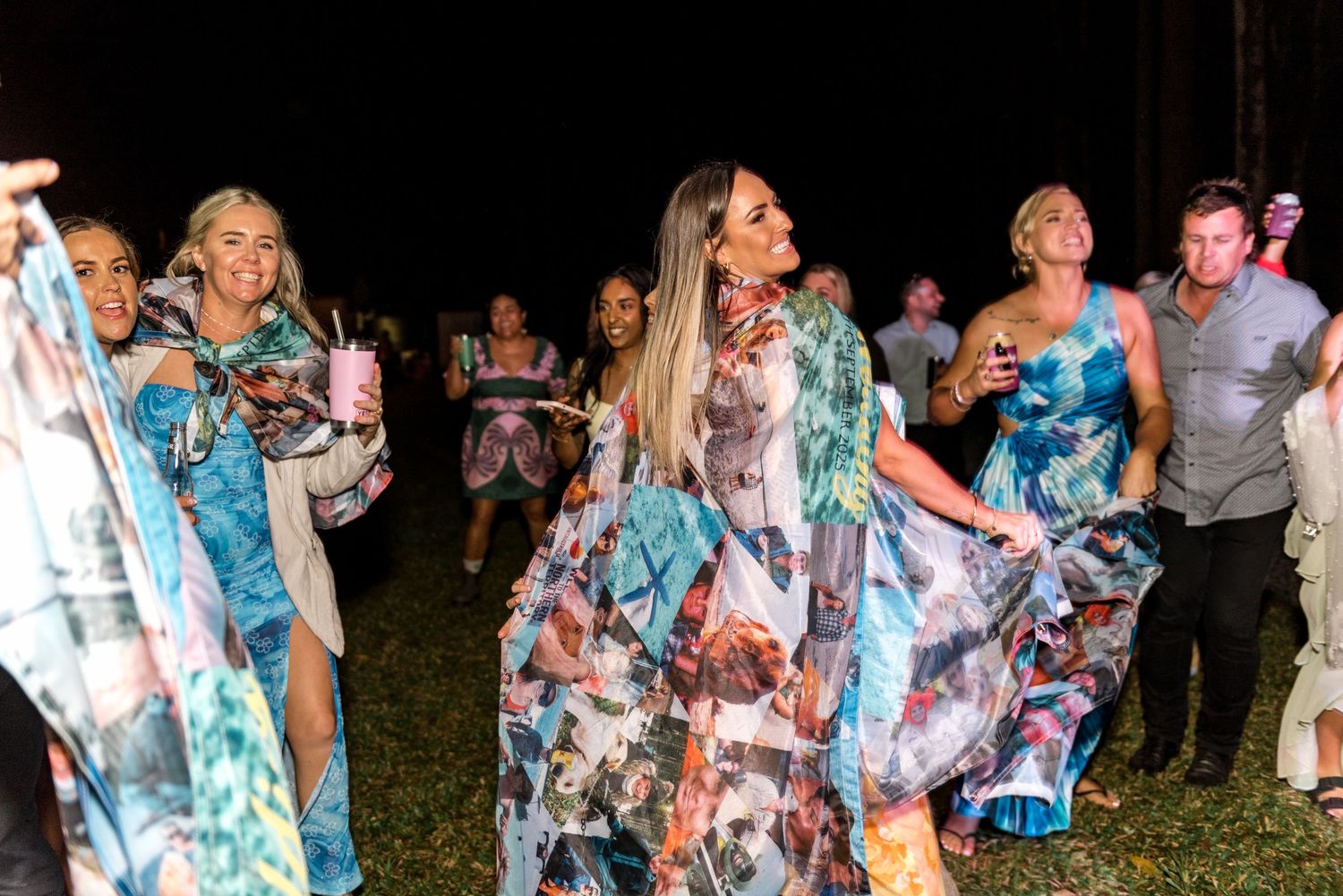 Group of women in colorful floral dresses dancing joyfully at an outdoor night event.