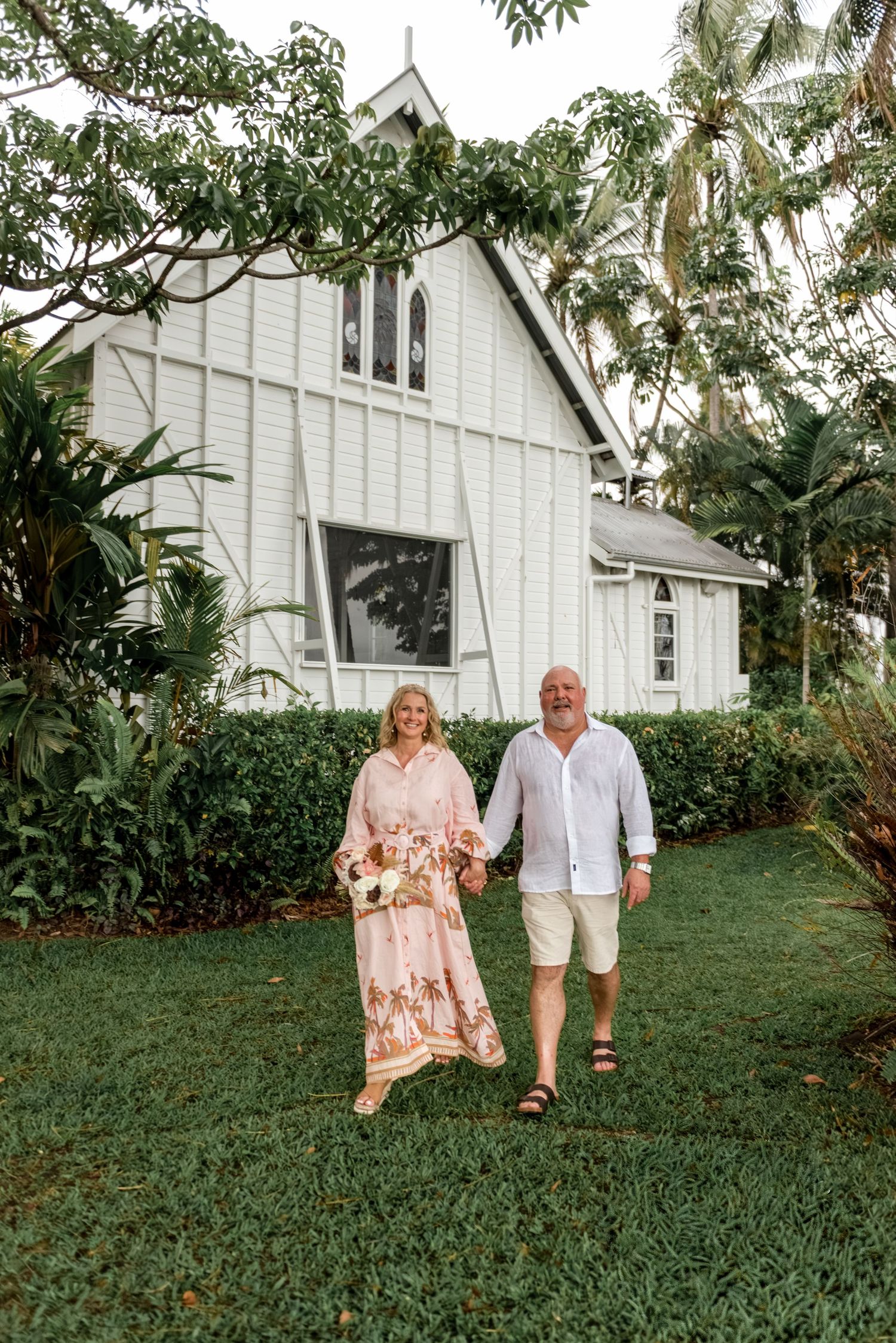 Happy couple walking hand in hand on green lawn in front of a white heritage church.