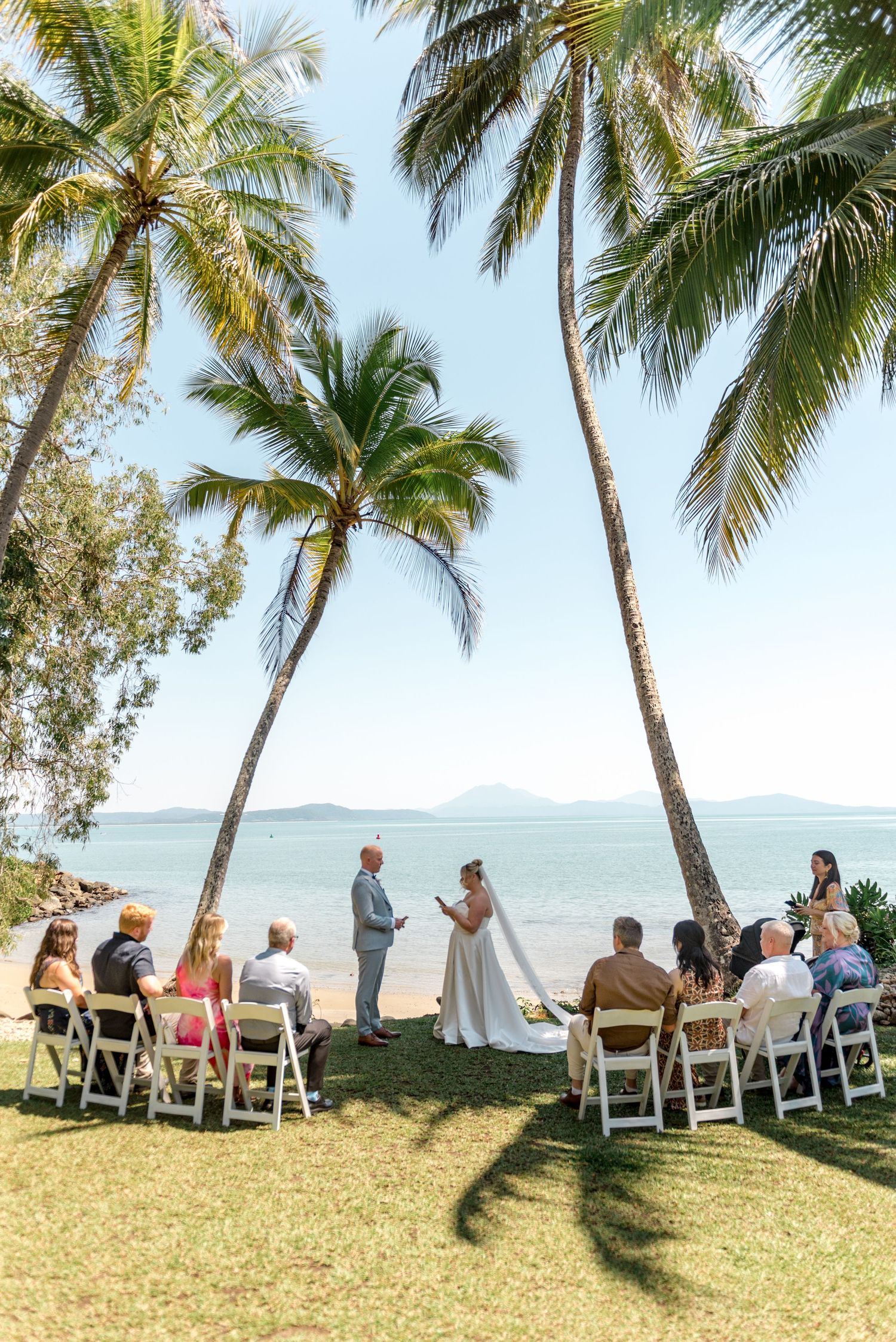 Small intimate beach wedding ceremony with guests seated on white chairs under tropical palm trees.