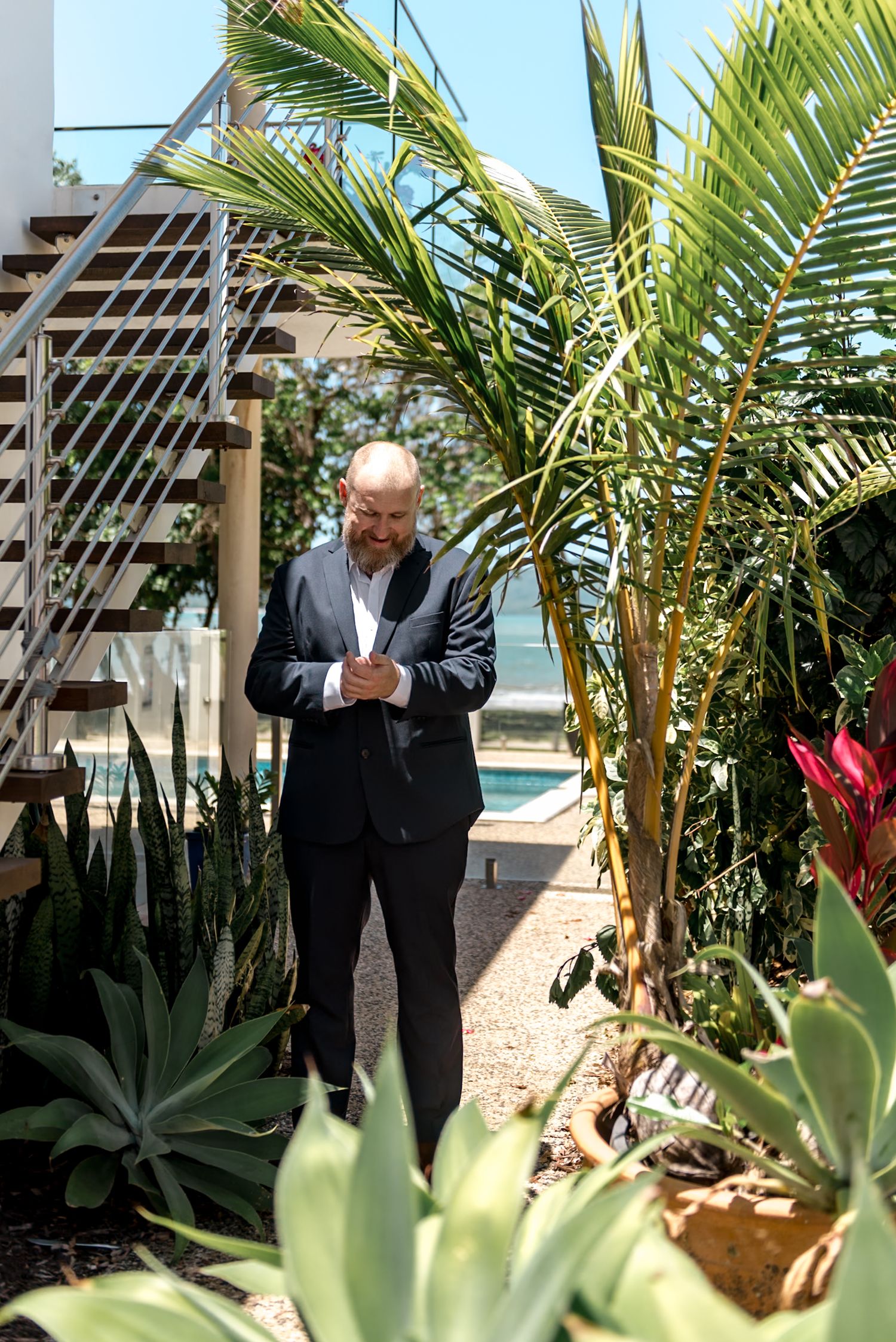 Bald man in a dark suit standing among tropical plants near a beachside staircase.