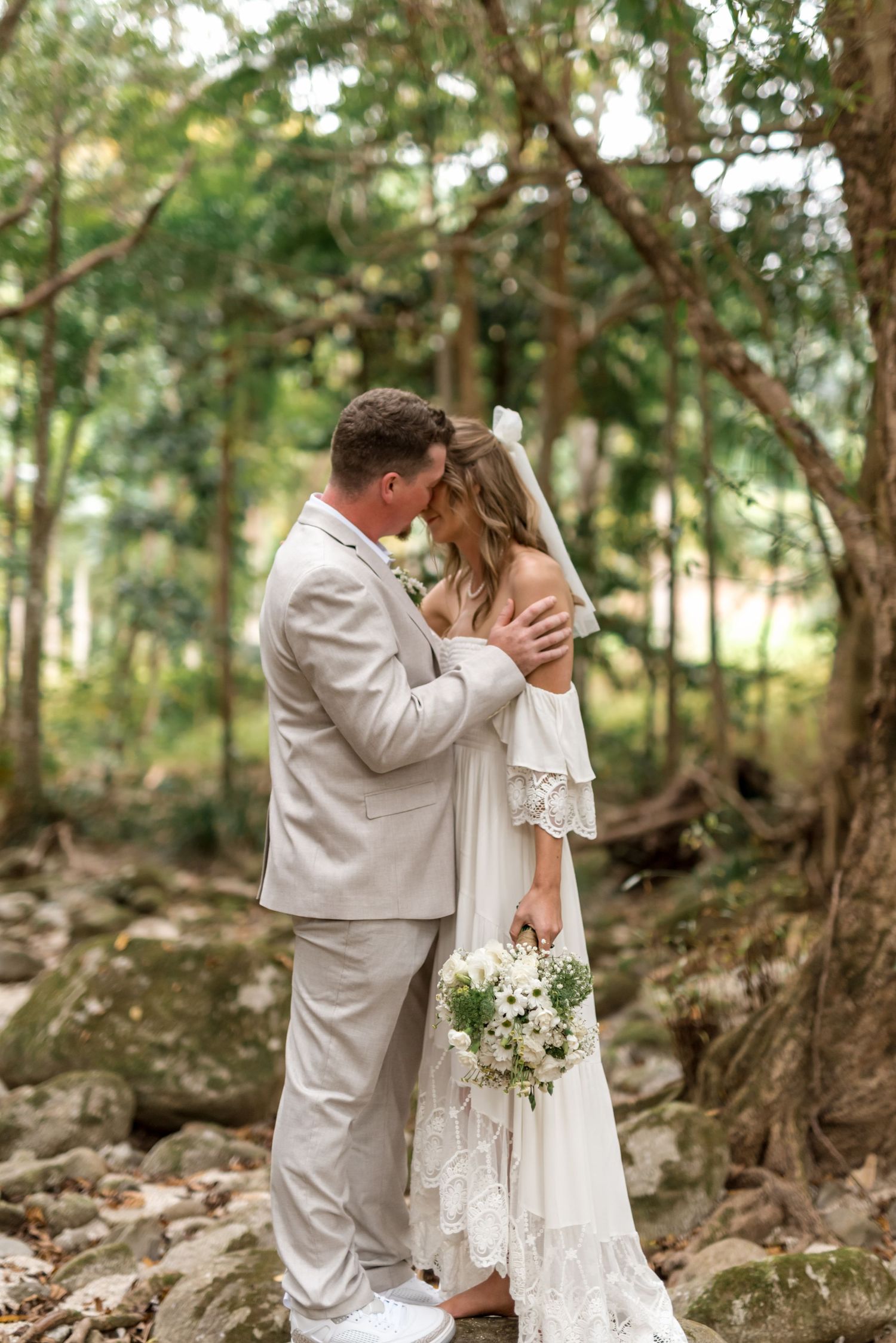 Couple sharing romantic moment in lush forest, bride in white boho lace dress holding bouquet, groom in light grey suit.