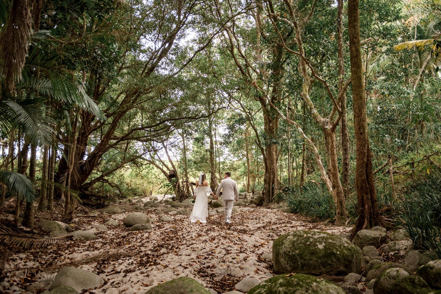 Newlywed couple walking hand-in-hand through sunlit forest with rocks and fallen leaves on the ground.