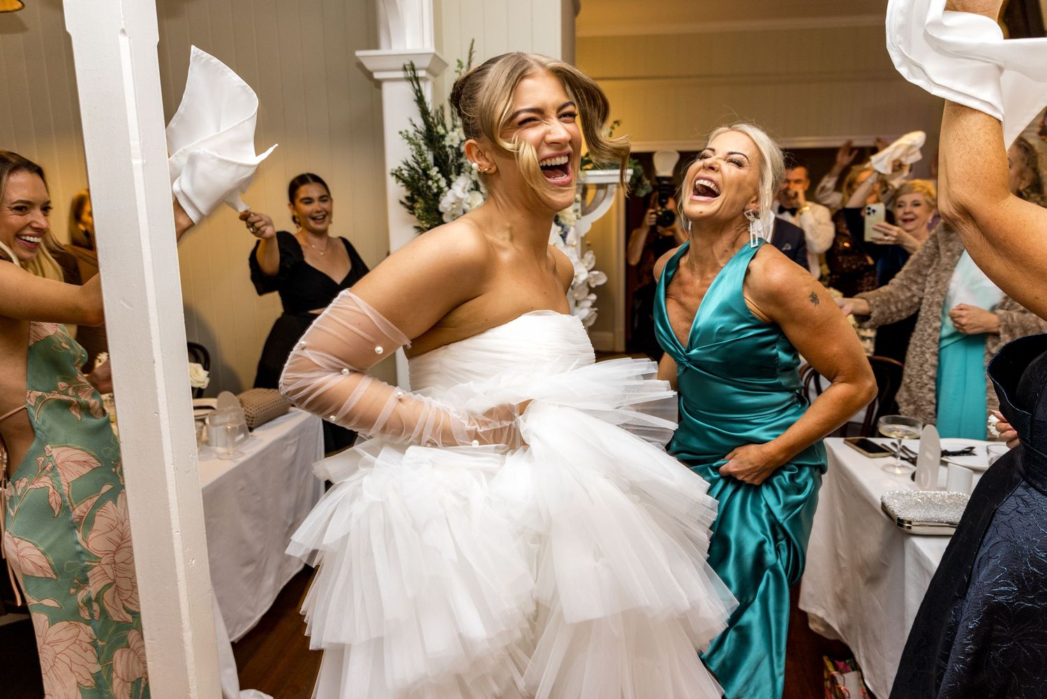 Laughing bride in white tulle gown celebrating with smiling guests at wedding reception.