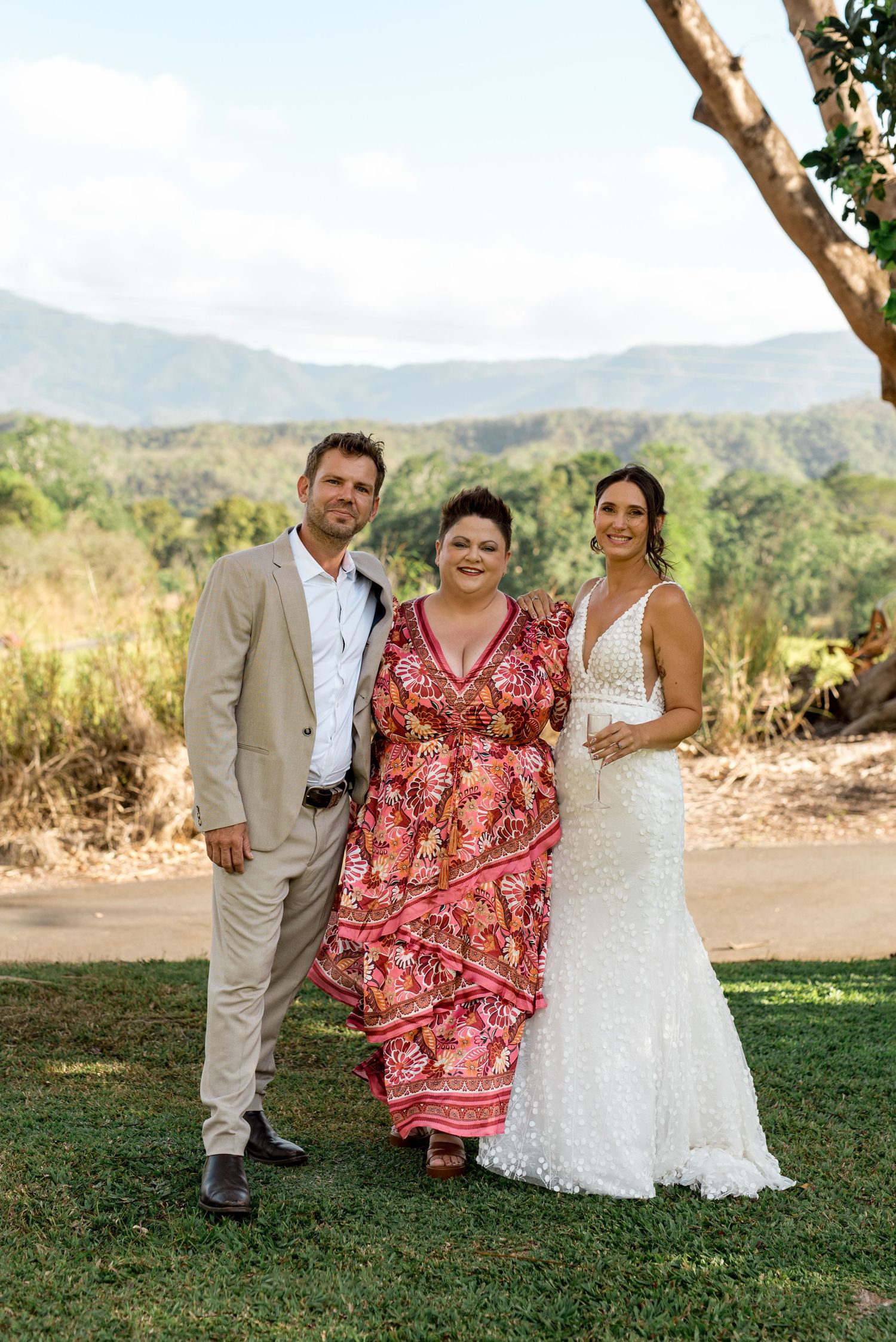 Bride in lace gown and groom in tan suit pose with guest against mountain backdrop.