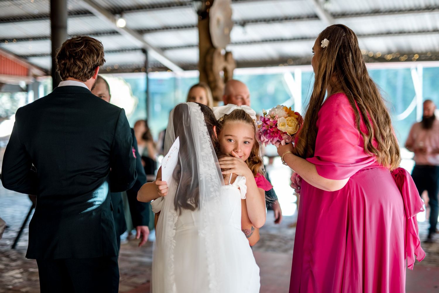 Flower girl in white dress hugs bride from behind as bridesmaid in pink assists at wedding reception.