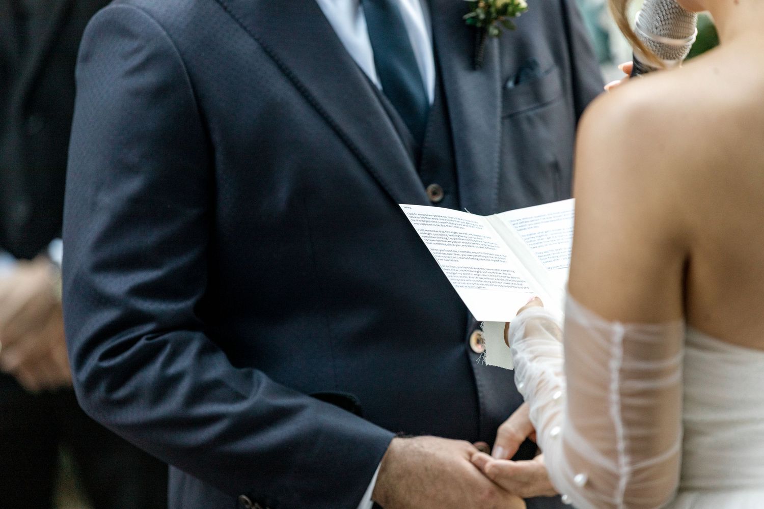 Groom reading wedding vows from paper while holding brides hands during ceremony.
