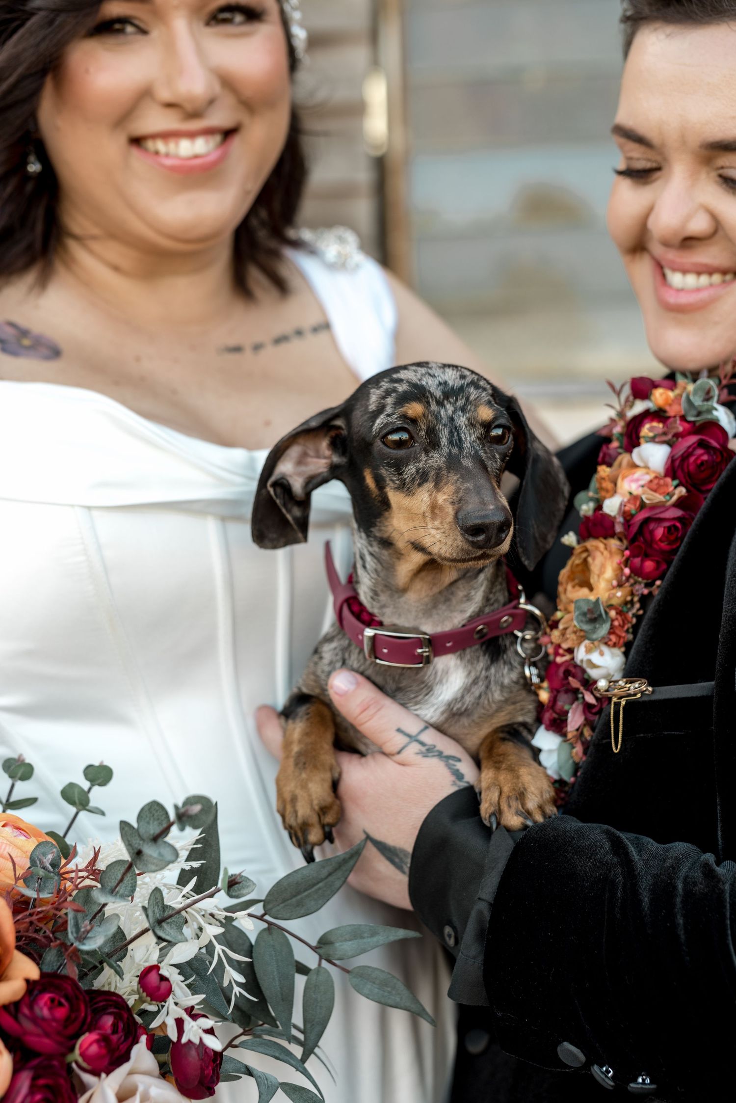Two brides smile holding an adorable dachshund puppy wearing a floral collar at their wedding.