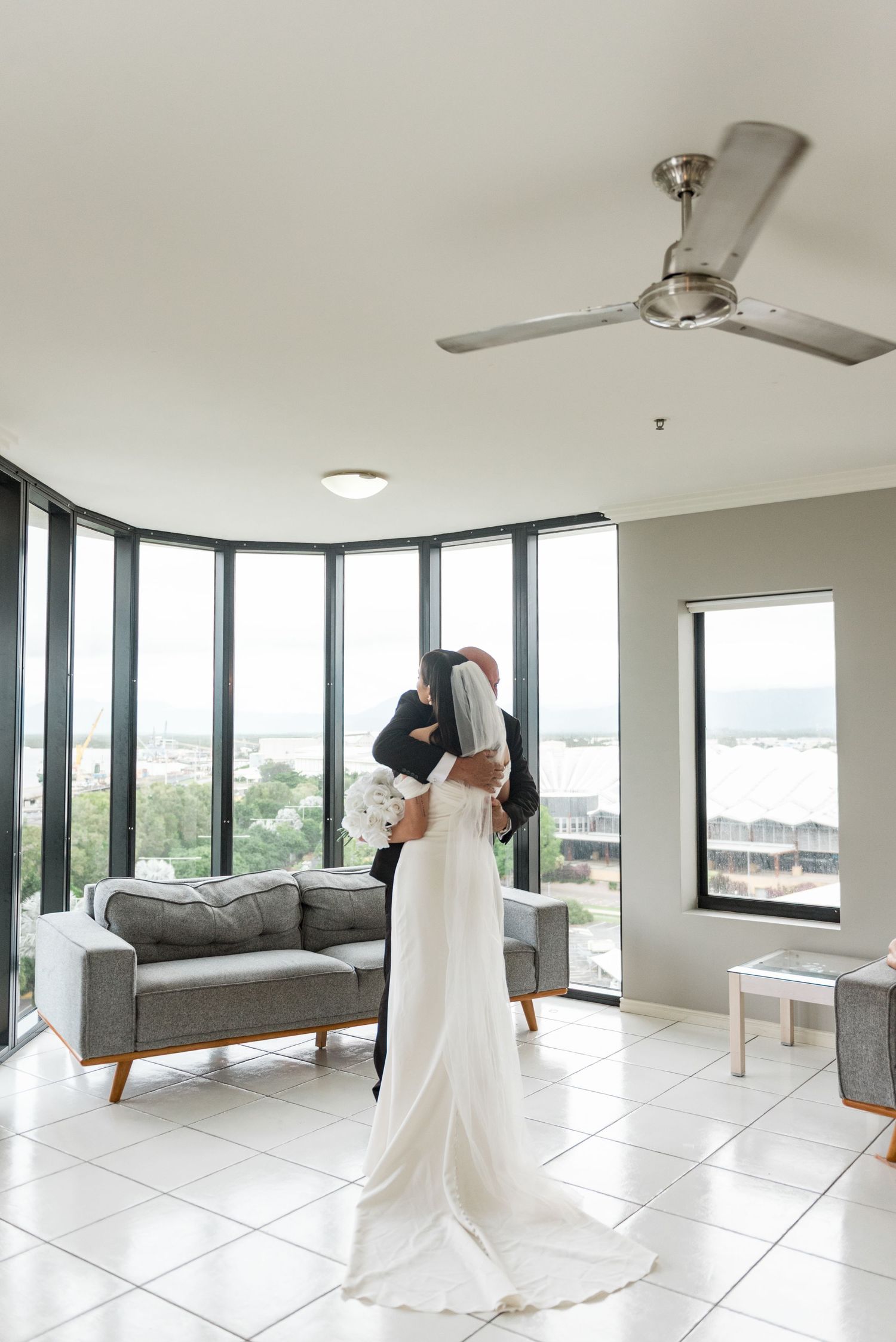 Bride and groom share a romantic embrace in a modern room with floor-to-ceiling windows and natural light.